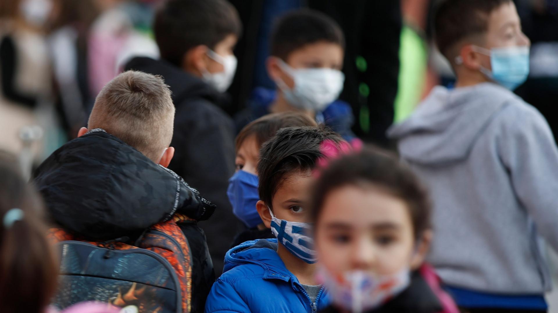A group of young students are shown wearing face masks and back packs as they wait in liine.