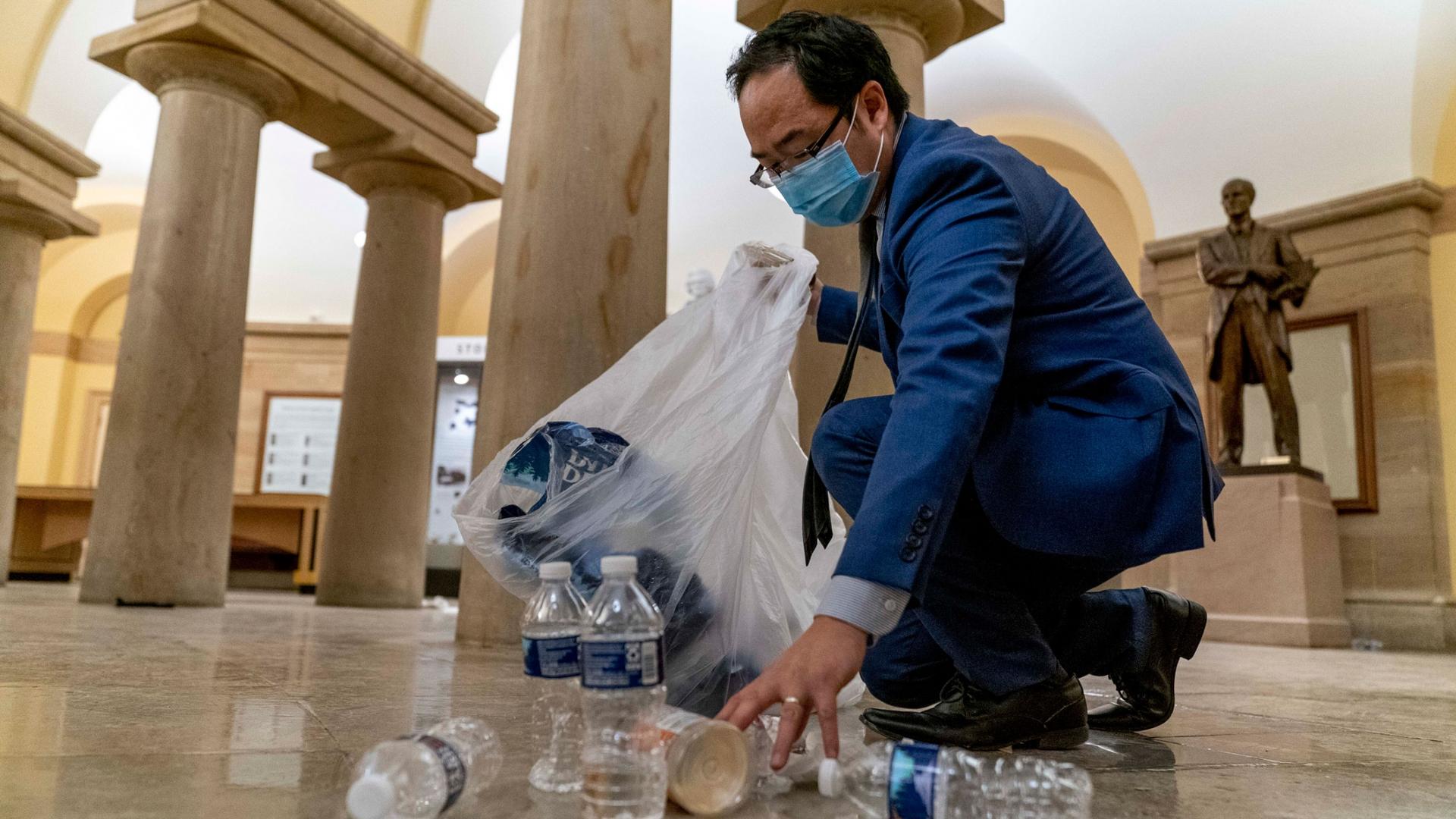 Rep. Andy Kim is shown kneeling and with a clear trash back helping clean up debris and trash strewn across the floor.