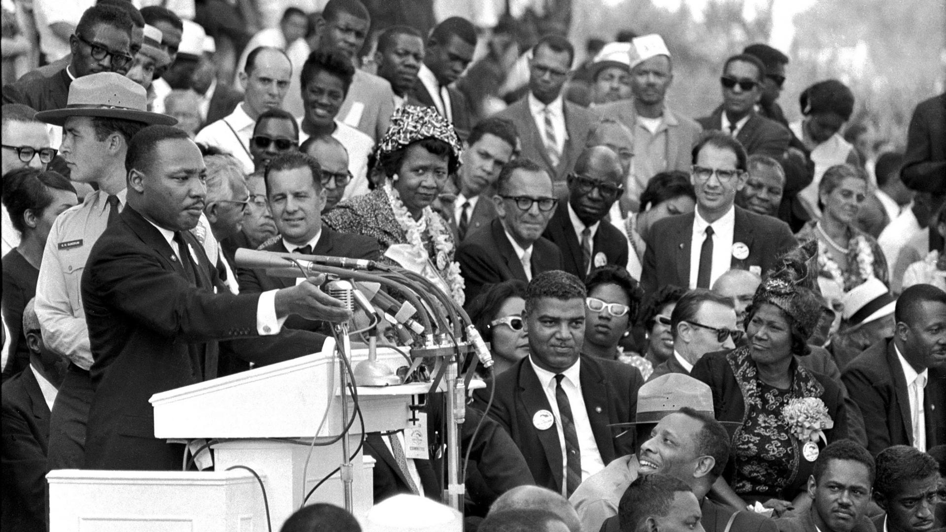 Rev. Dr. Martin Luther King Jr. is shown in a black and white photograph standing at a podium with several microphones and a crowd of people bedhind him.