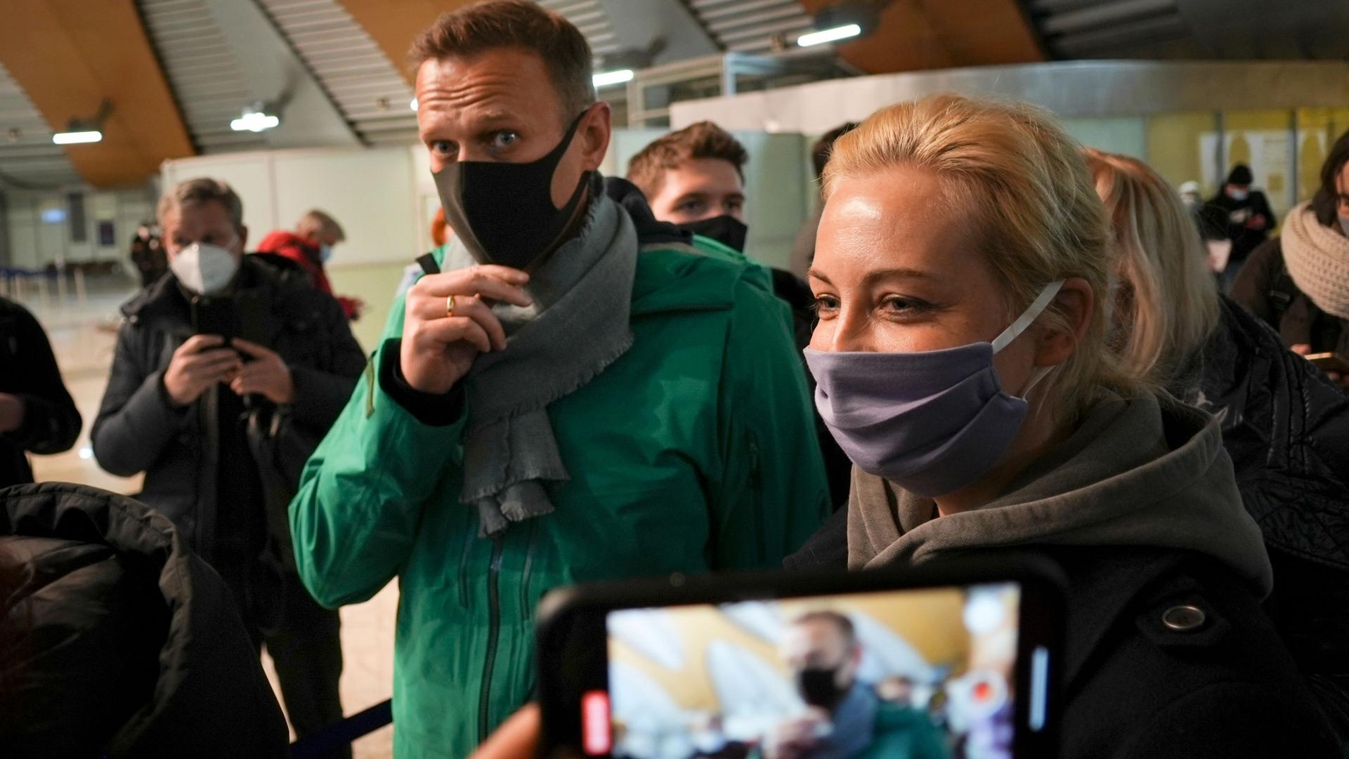 Alexei Navalny and his wife Yuliastand are shown wearing face masks and standing next to each other with a mobile phone also showing them in the near ground.