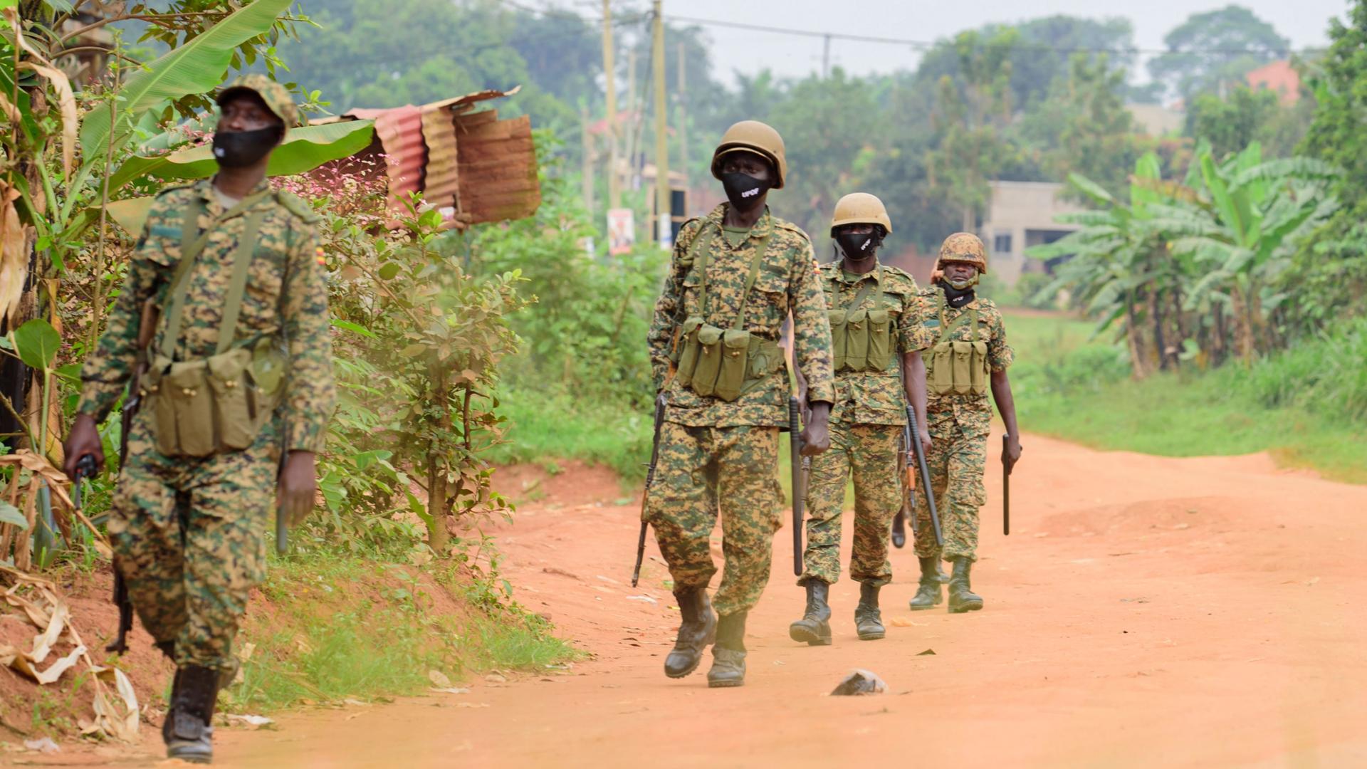 Four soldiers are shown wearing military fatigues and helmets while carrying weapons and walking in a dirt road.