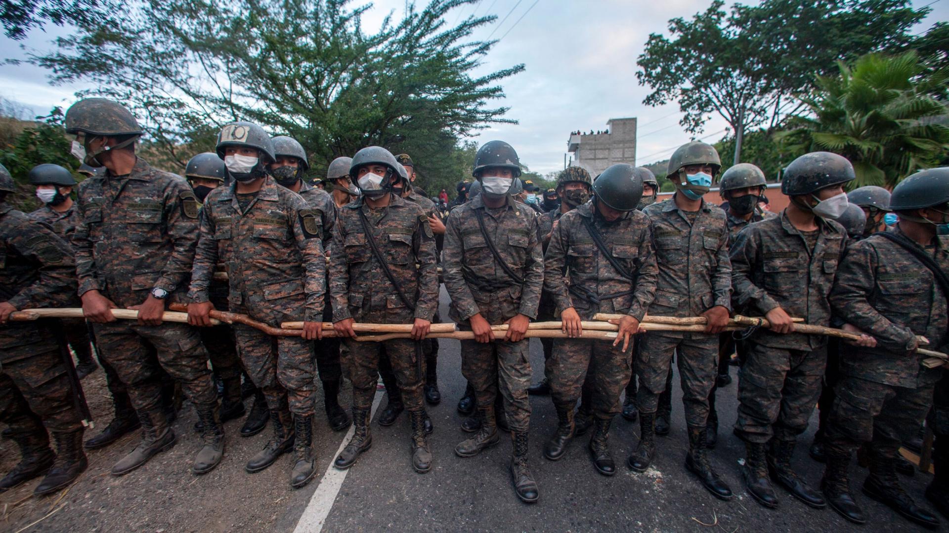A line of security authorities are shown wearing fatigues and helmets and holding wooden poles.