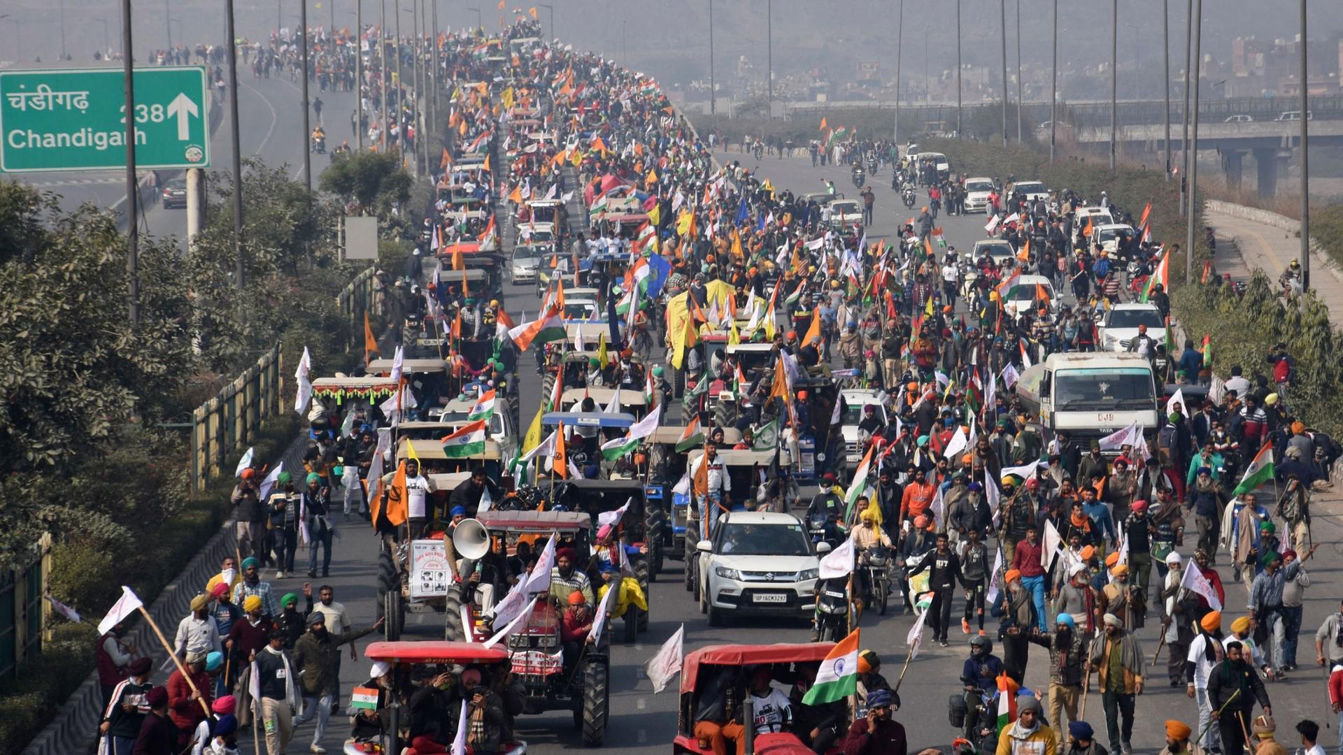 A multiple lane road is shown filled with protesters driving tractors and other vehicles with thousands of people shown all over the road.