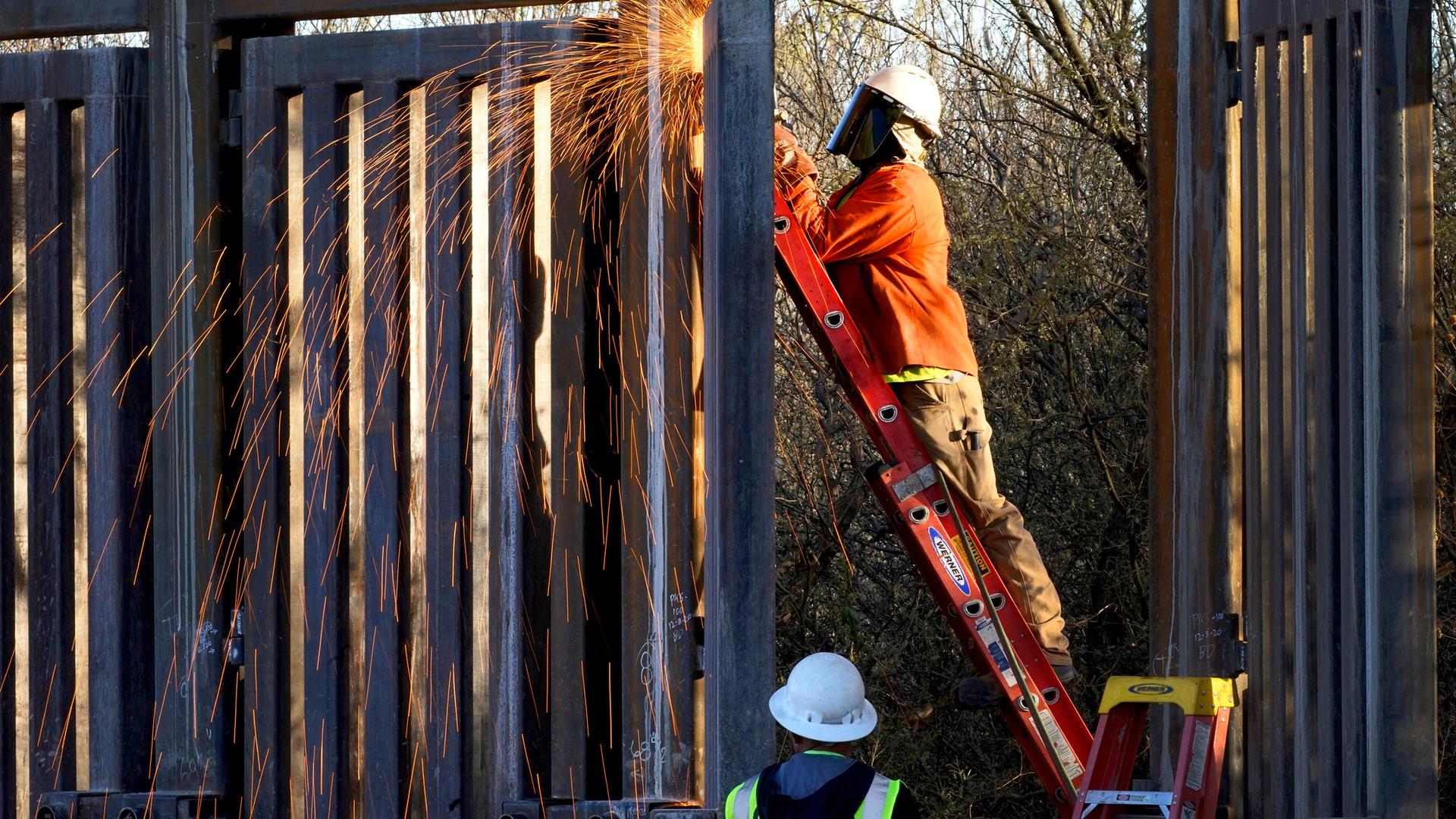 A man is shown standing on a ladder and using a machine causing sparks to fly in the construction of a border wall.