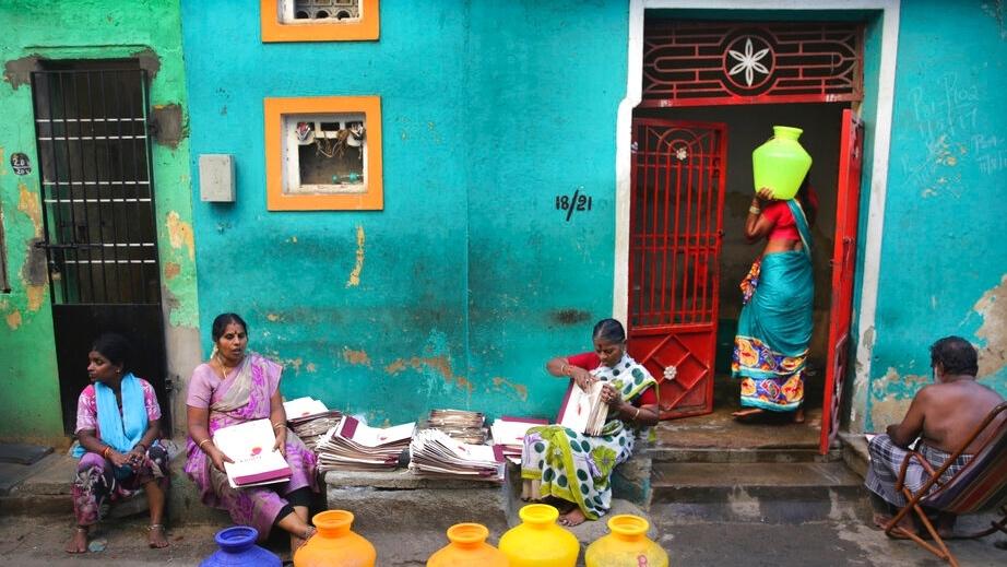 Several women in colorful saris sit and stand with bright yellow drinking vessels in front of a blue and green painted building. 