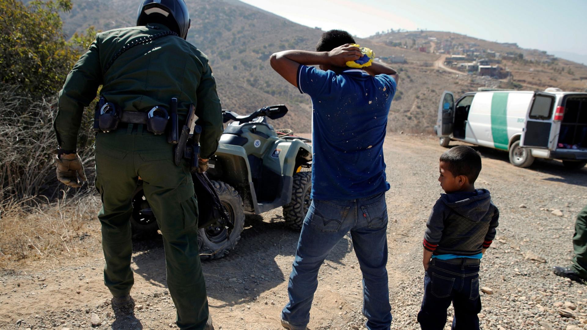 A man is shown with his hands behind his head as he is apprehended by a US border agent while a small boy is standing nearby.
