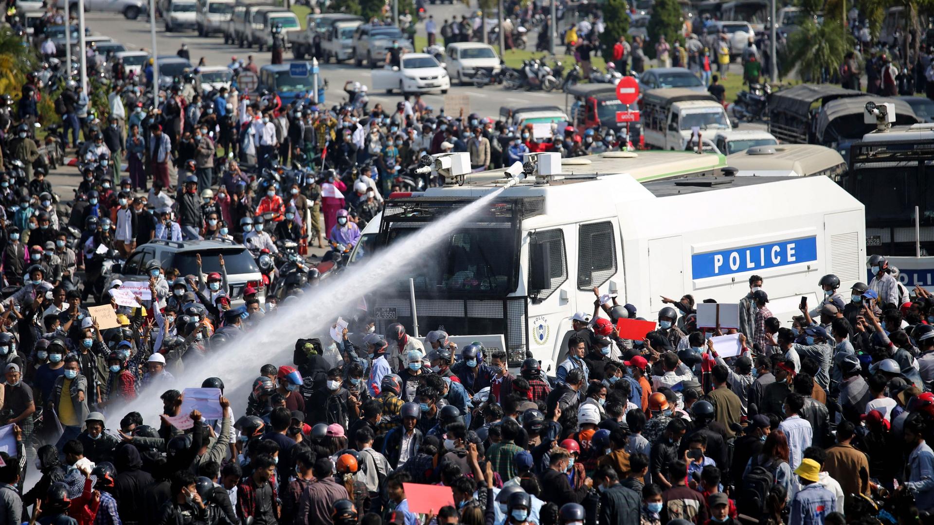 A large crowd of people are shown surrounding a police truck which is shooting a water cannon into the crowd.