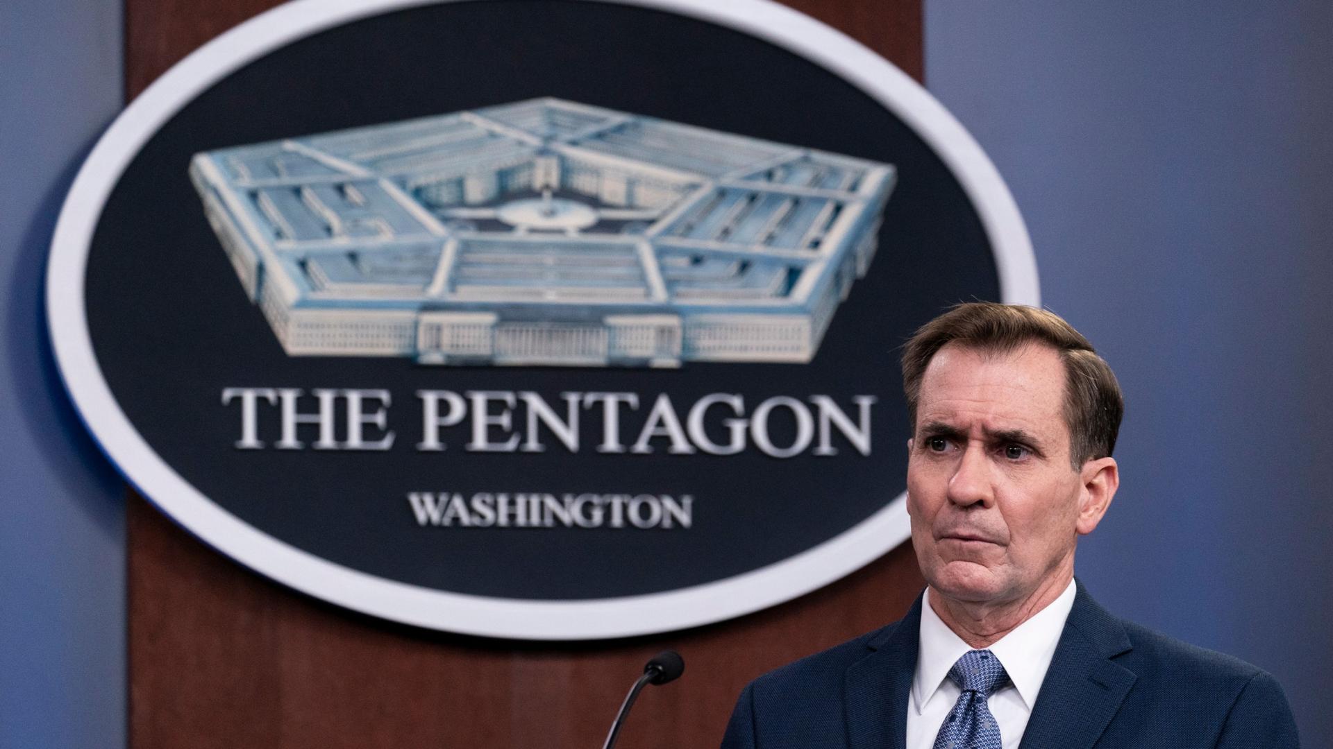 Pentagon spokesman John Kirby is shown wearing a dark blue suit and standing behind a microphone with the logo for the Pentagon behind him.