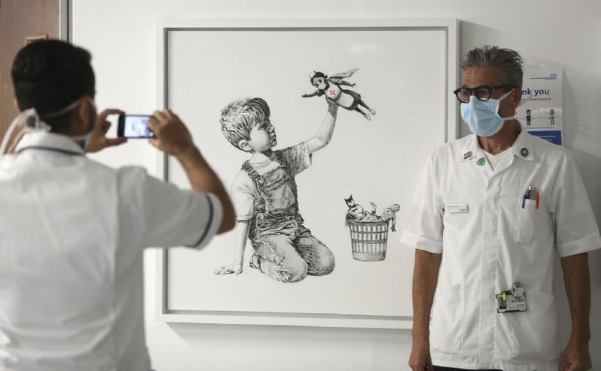 A member of a hospital staff is shown standing next to a Banksy artwork while getting their photograph taken.