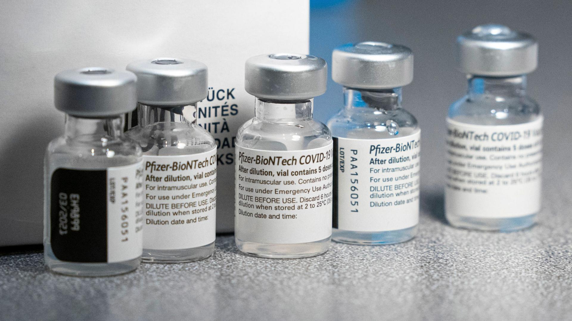 Five clear-glass vials are shown with labels showing that they are the Pfizer-BioNTech COVID-19 vaccine.