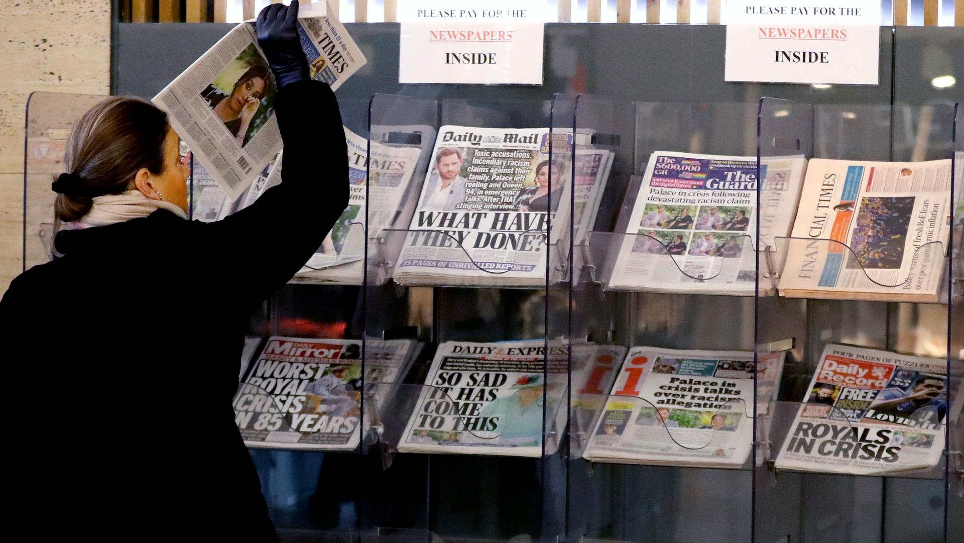 A woman is shown wearing a dark coat and pulling a newspaper out of a stand in a row of several other newspapers.