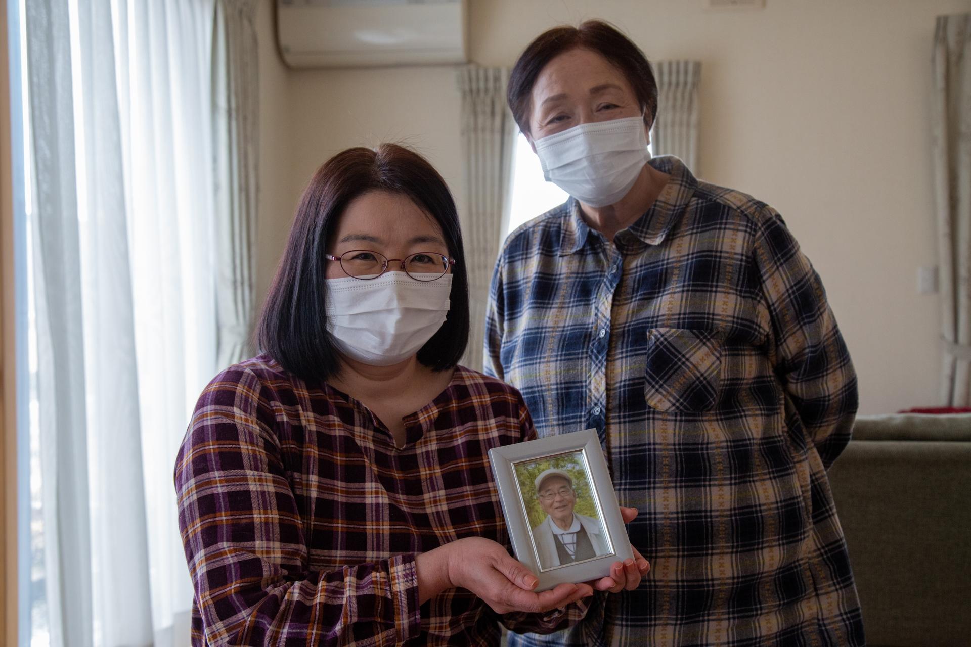 Two women are shown next two each other, both wearing medical face masks and one holding a small framed photograph.