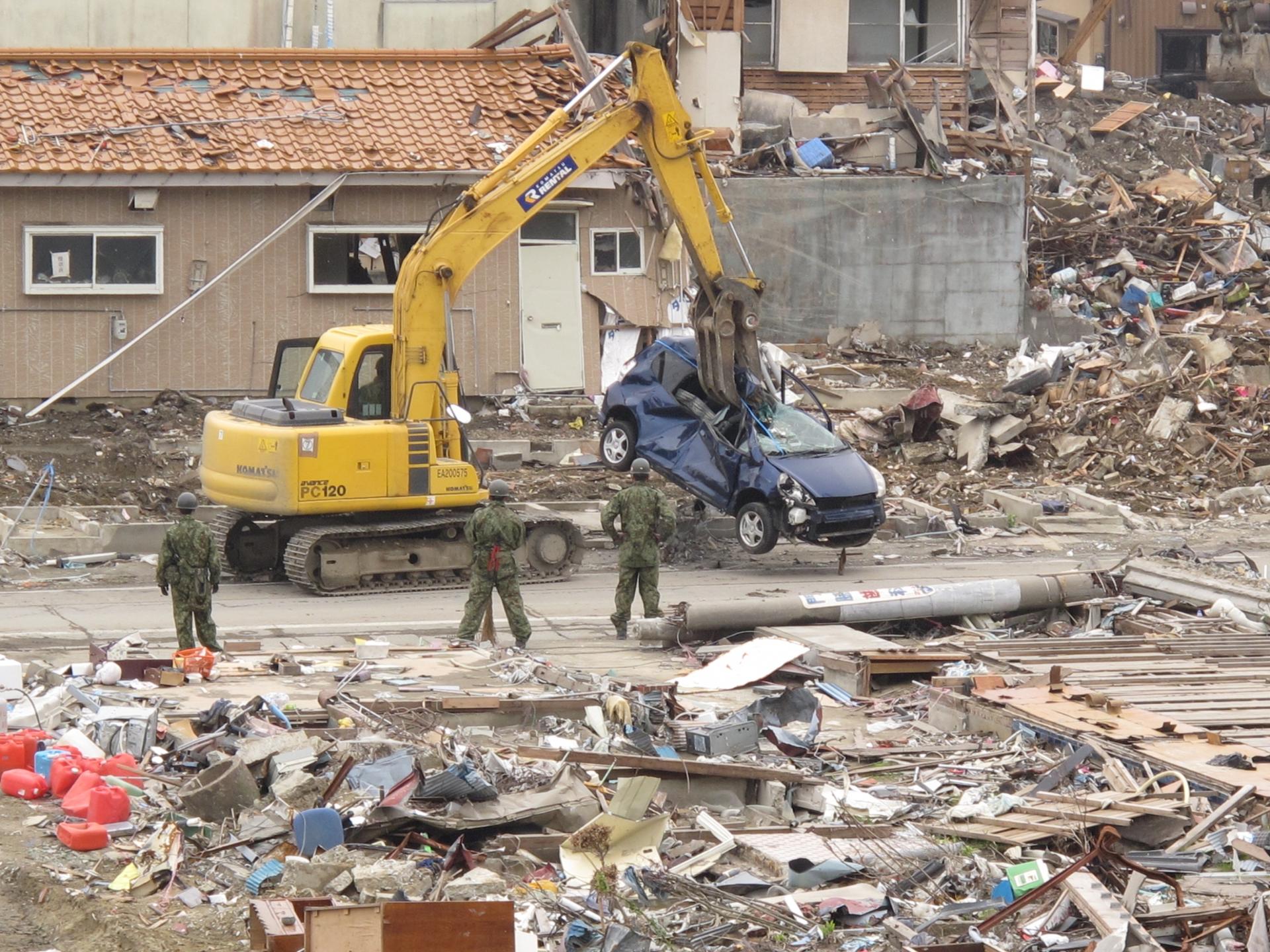 A yellow crane with a long arm is shown lifting up a destroyed blue car with damaged buildings all over.
