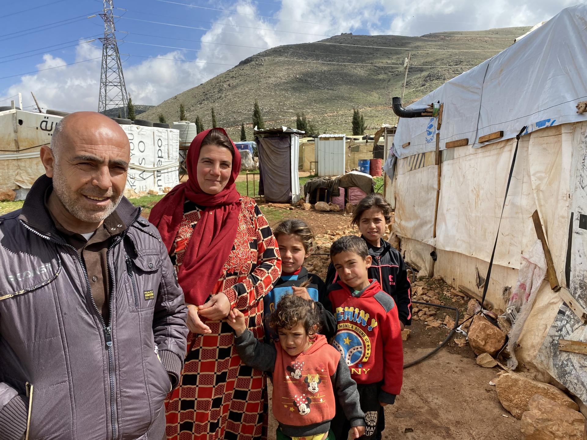Ali Hamoud stands in front of his tent with his family. The mountains behind mark the Syrian border. 
