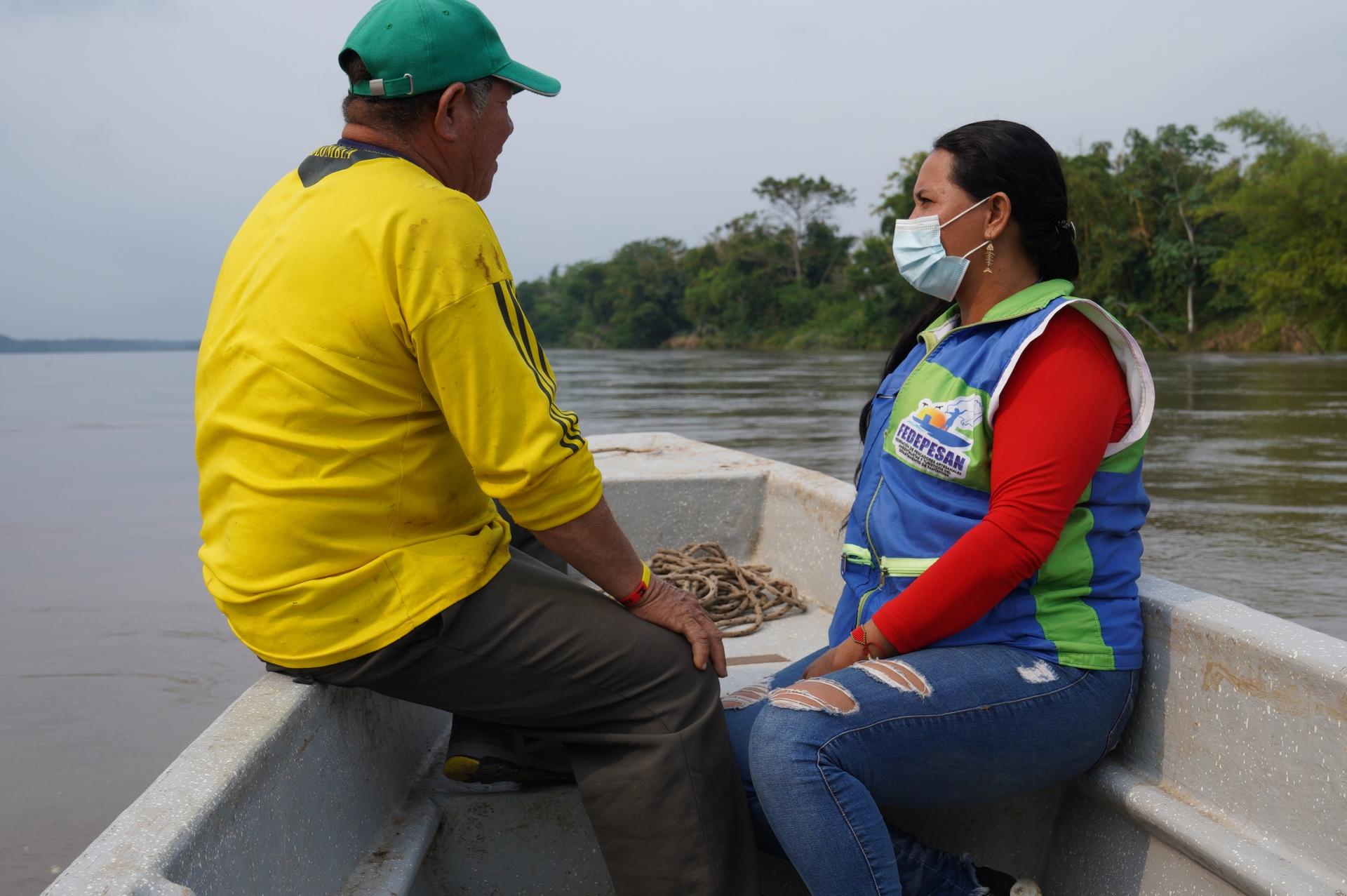 A man wearing a yellow shirt and green cap and a woman wearing a blue vest with a mask ride a boat on a river. 