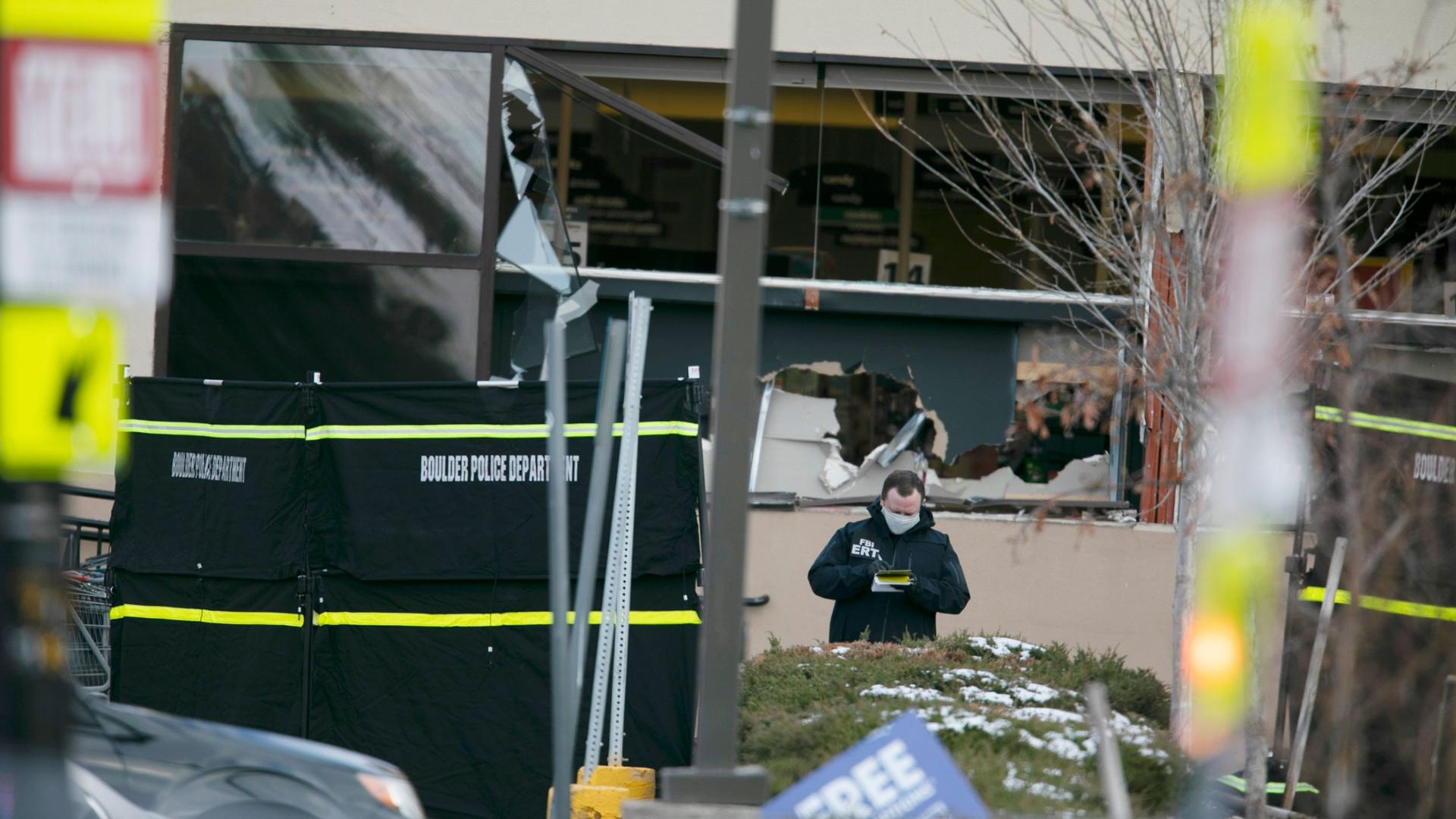 A police officer is shown wearing a face mask amidst broken glass and other remnants of a shooting with Boulder Police baracades in the nearground.