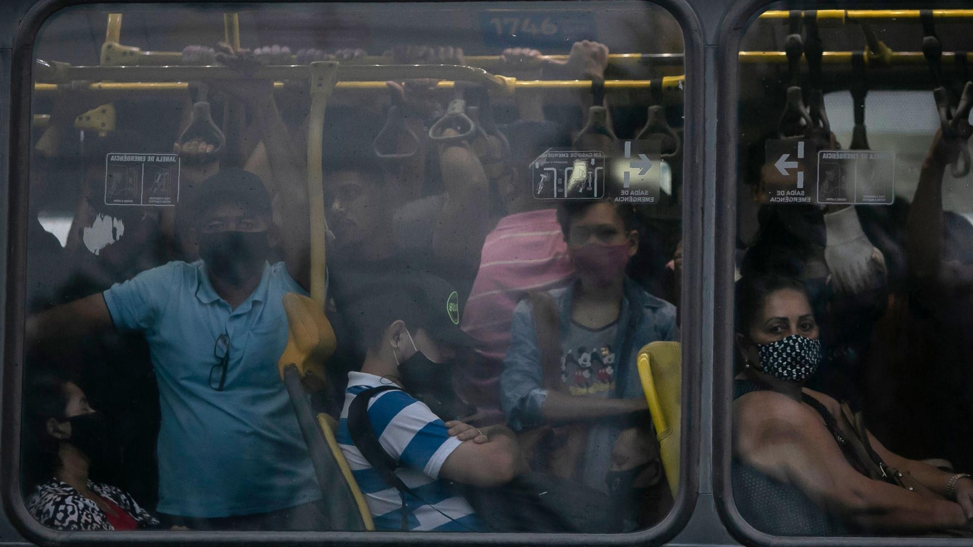 A crowd of people are shown crammed onto a bus as shown through the window.