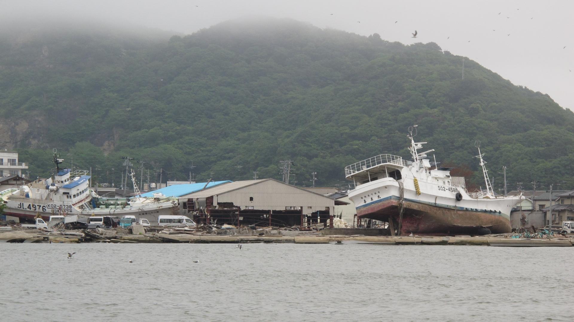 Several large boats are shown on their side and washed ashore following a tsunami as shown from across a body of water.
