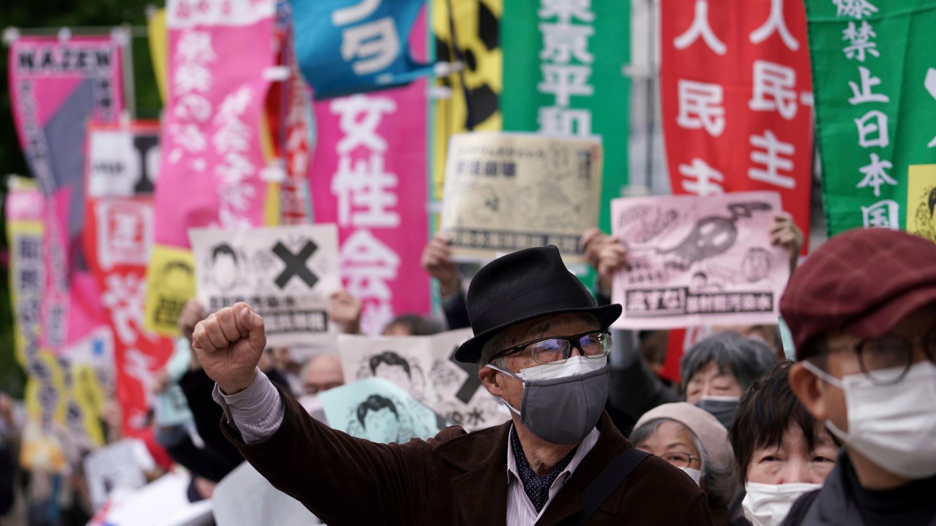 A man is shown among a crowd of protesters wearing a dark jacket, hat and face mask with his right hand in the air.