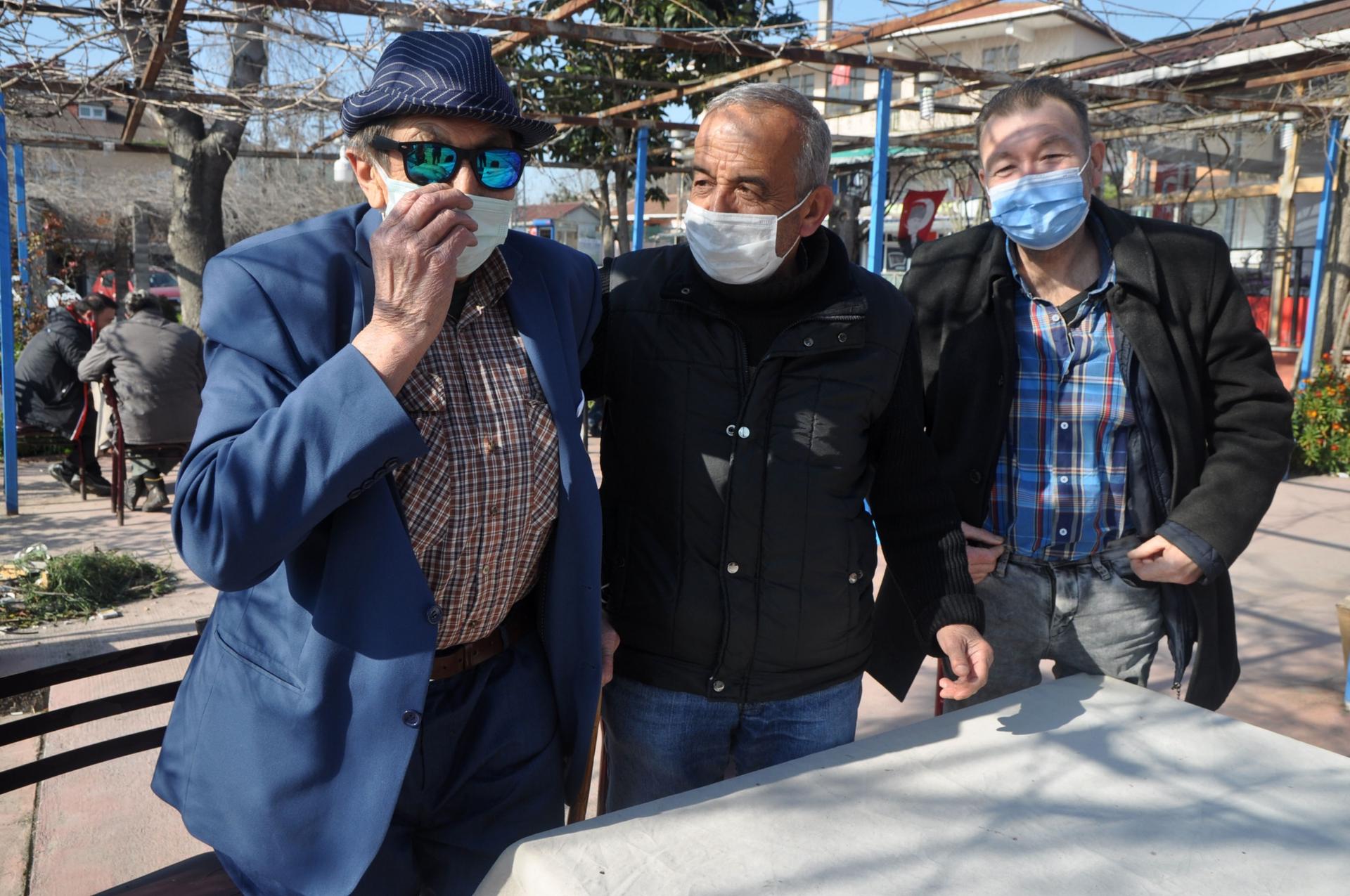 Three older men wearing blue and black clothing and face masks pose together in an outdoor tea garden. 