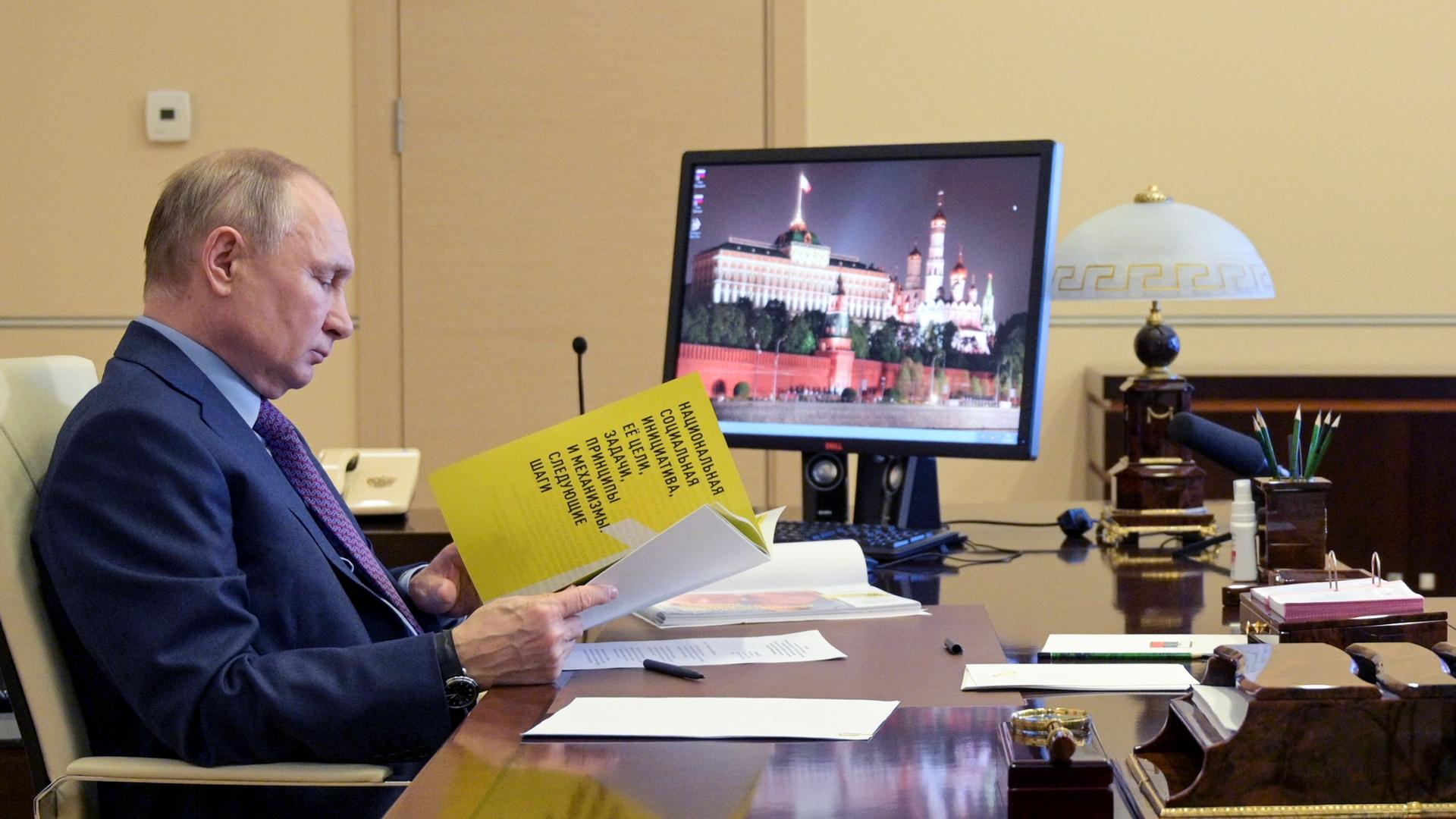 Russian President Vladimir Putin is shown sitting at a desk and looking down at a yellow document.