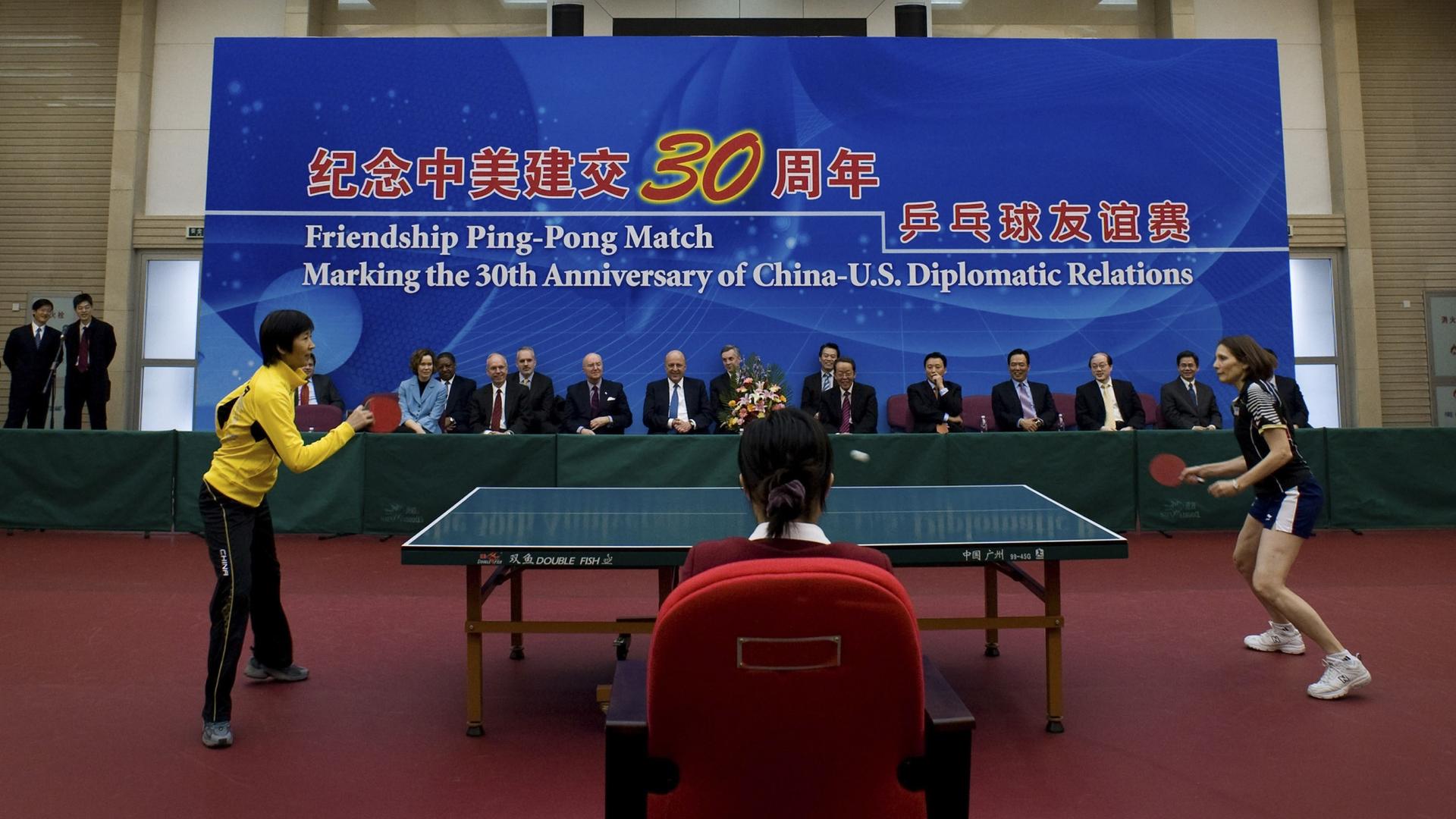 Table top tennis players compete in front of a large blue banner