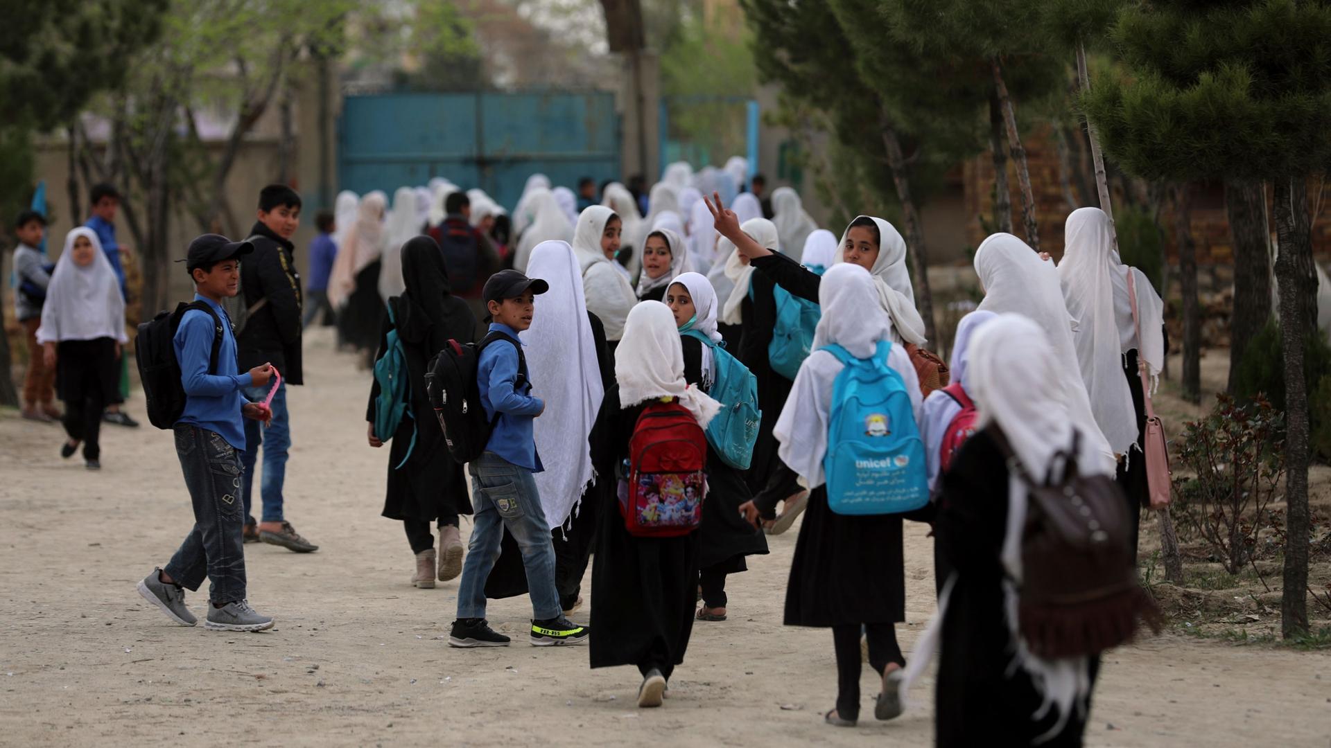 A crowd of young people are shown walking near a school with many girls wearing a white head covering.