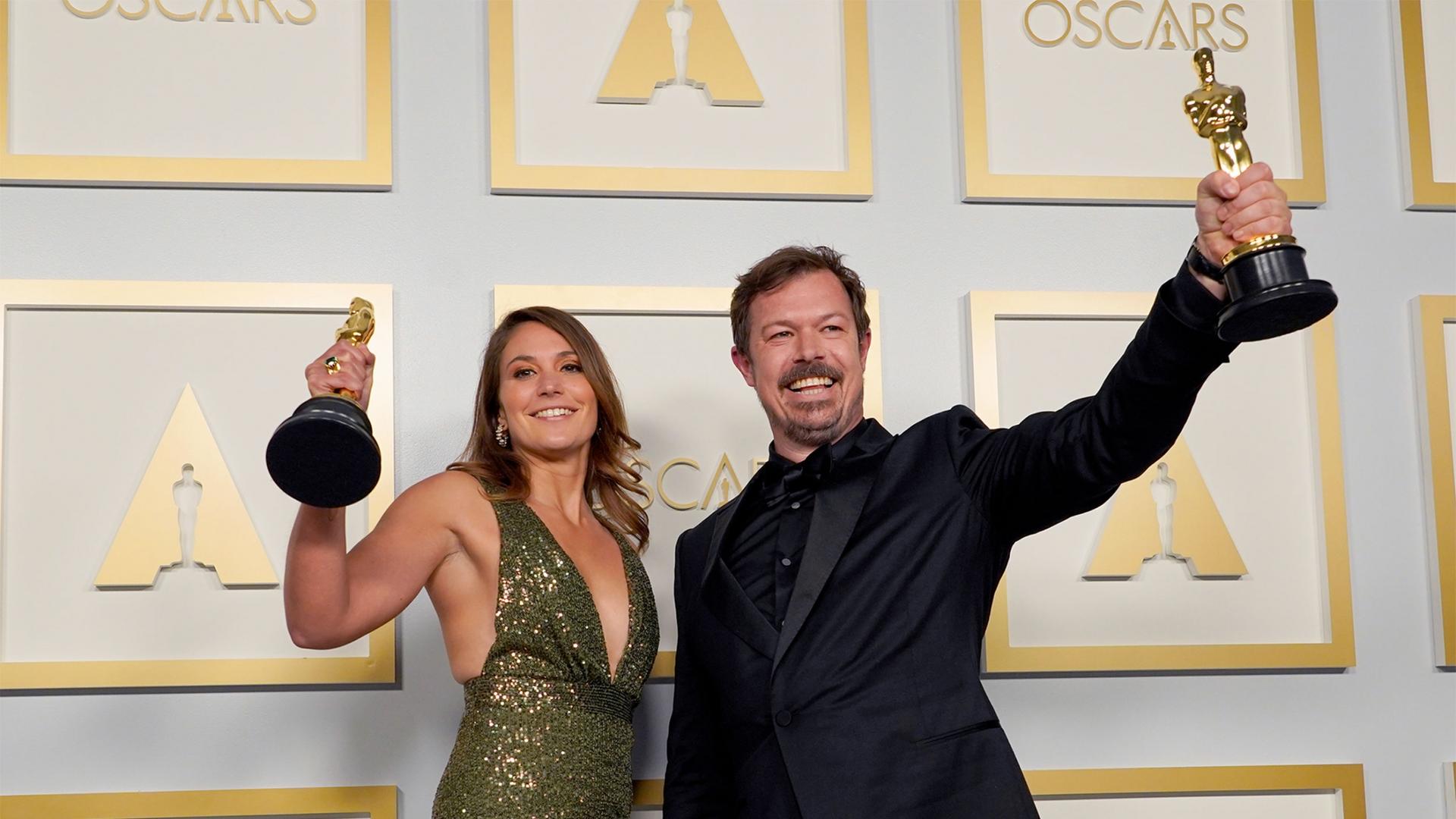 Woman in green dress and man in black suit hold up Oscars