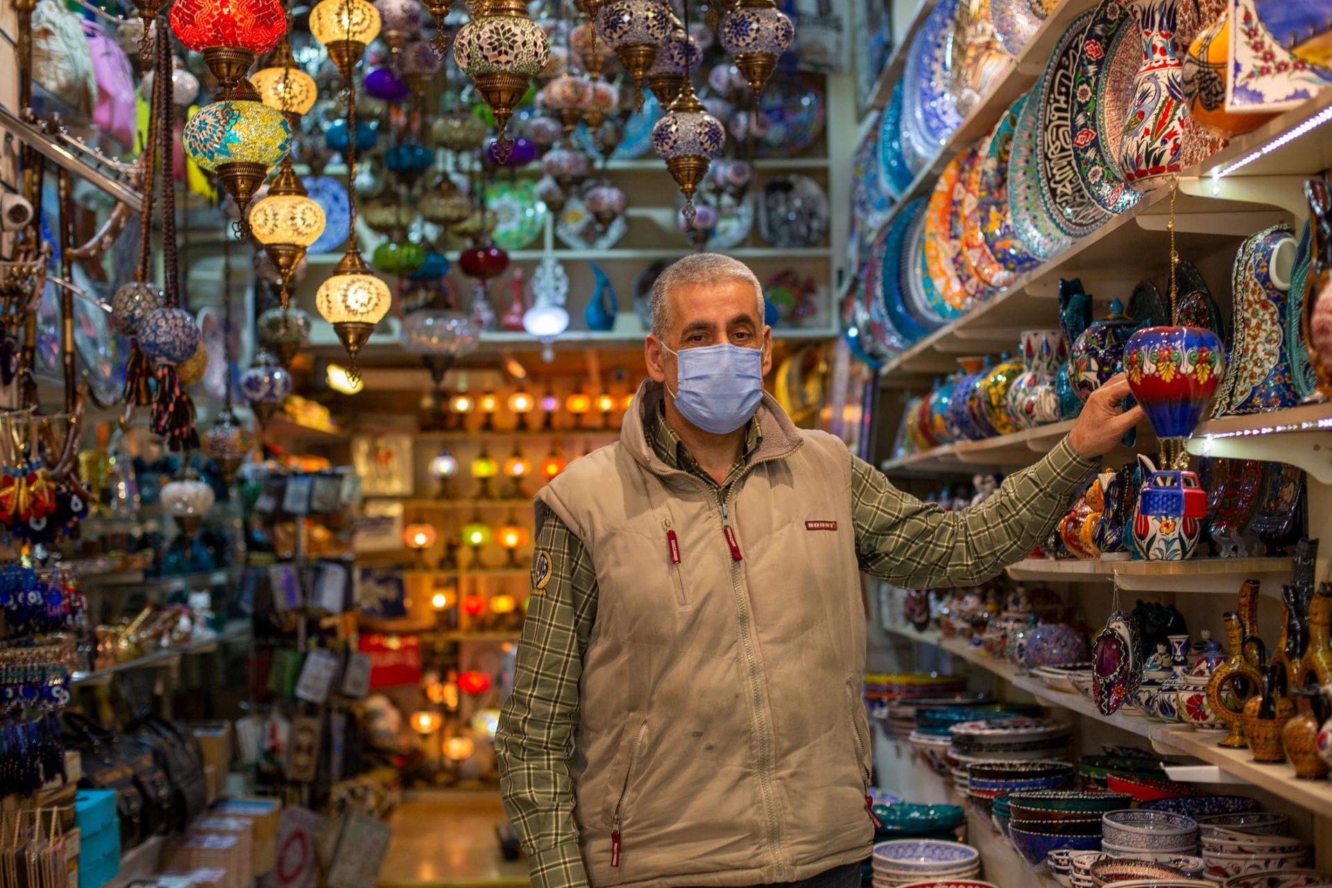 A man is shown wearing a face mask and khaki vest while standing in an aisle with colorful lamps.