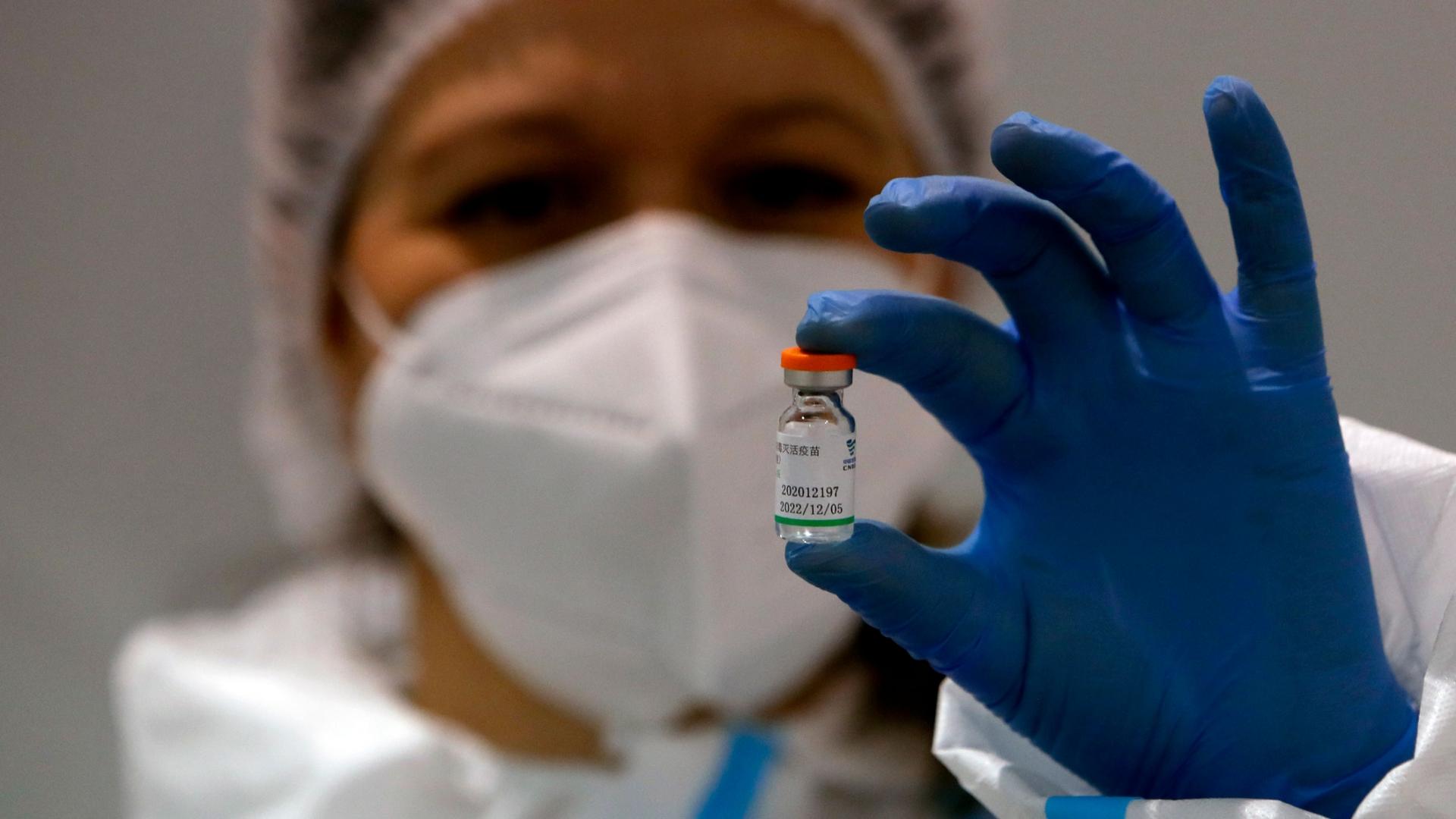 A person is shown wearing a protective medical mask and hair net while holding between their finfers a file of the Sinopharm's COVID-19 vaccine.