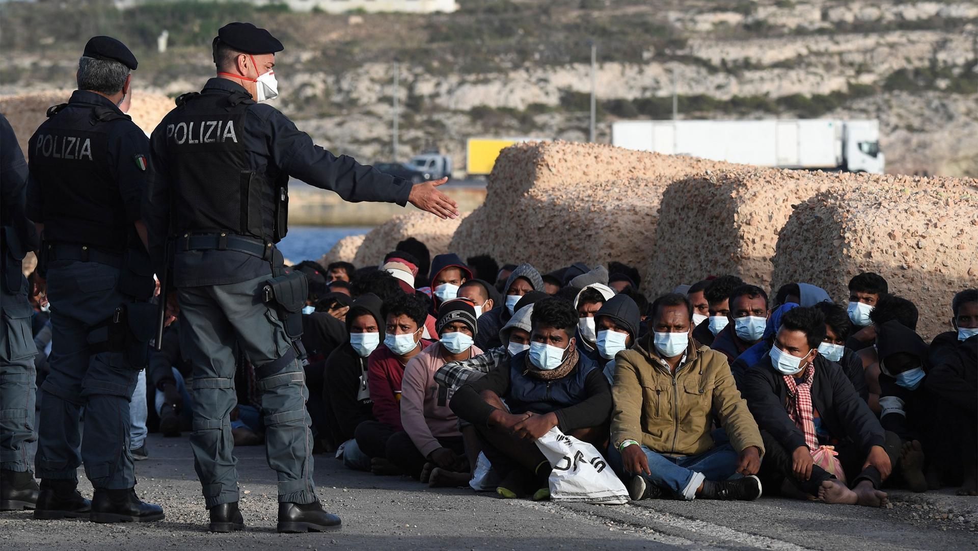 Migrants wearing face masks to curb the spread of COVID-19 sit at a pier as Italian police officers stand by.