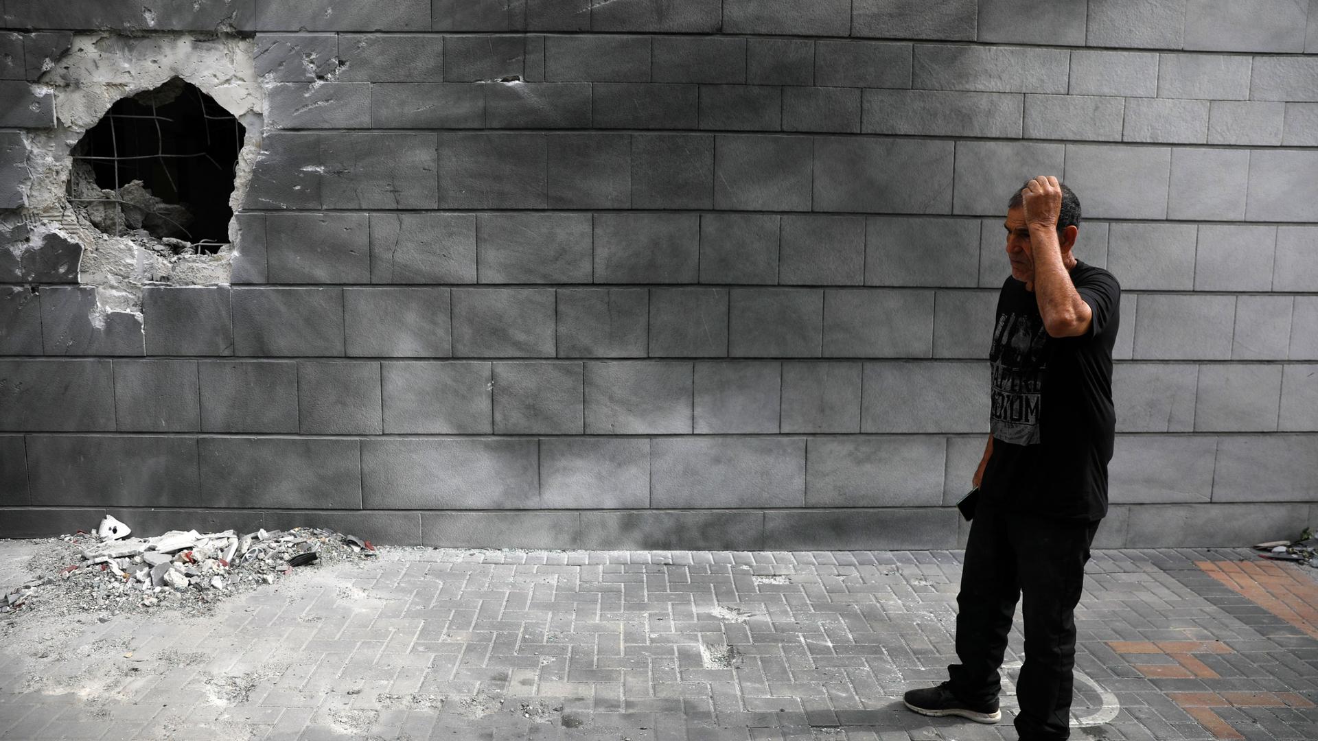 A man is show wearing a black t-shirt and pants while standing next to a cinder block wall with a large hole in it.