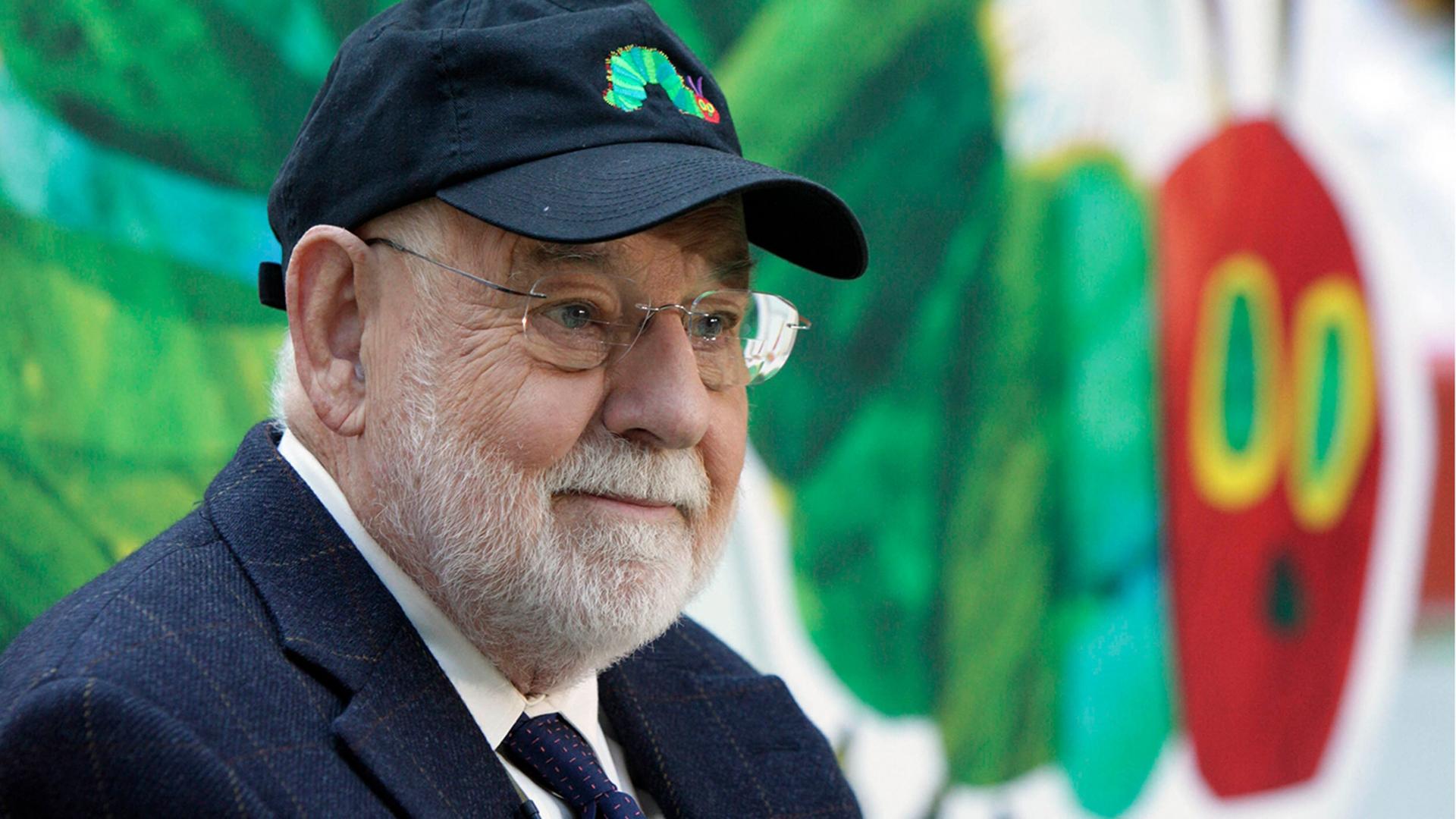 Author Eric Carle is shown with a white beard and warding a suit, tie and baseball hat.