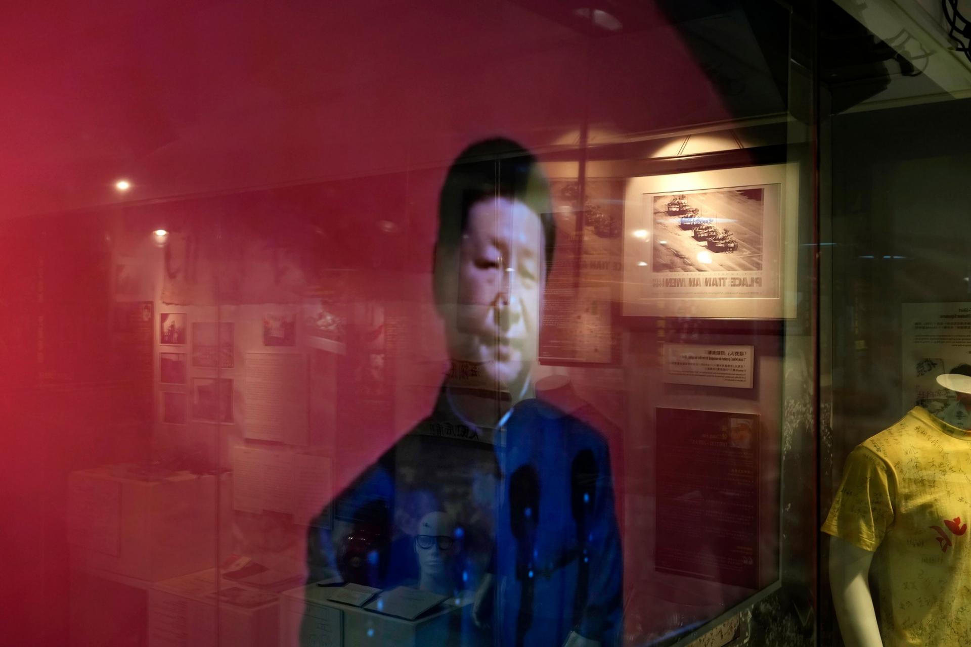 An image of China President Xi Jinping depicted on a red background is shown is shown through glass that's reflecting other Tiananmen Square artifacts.