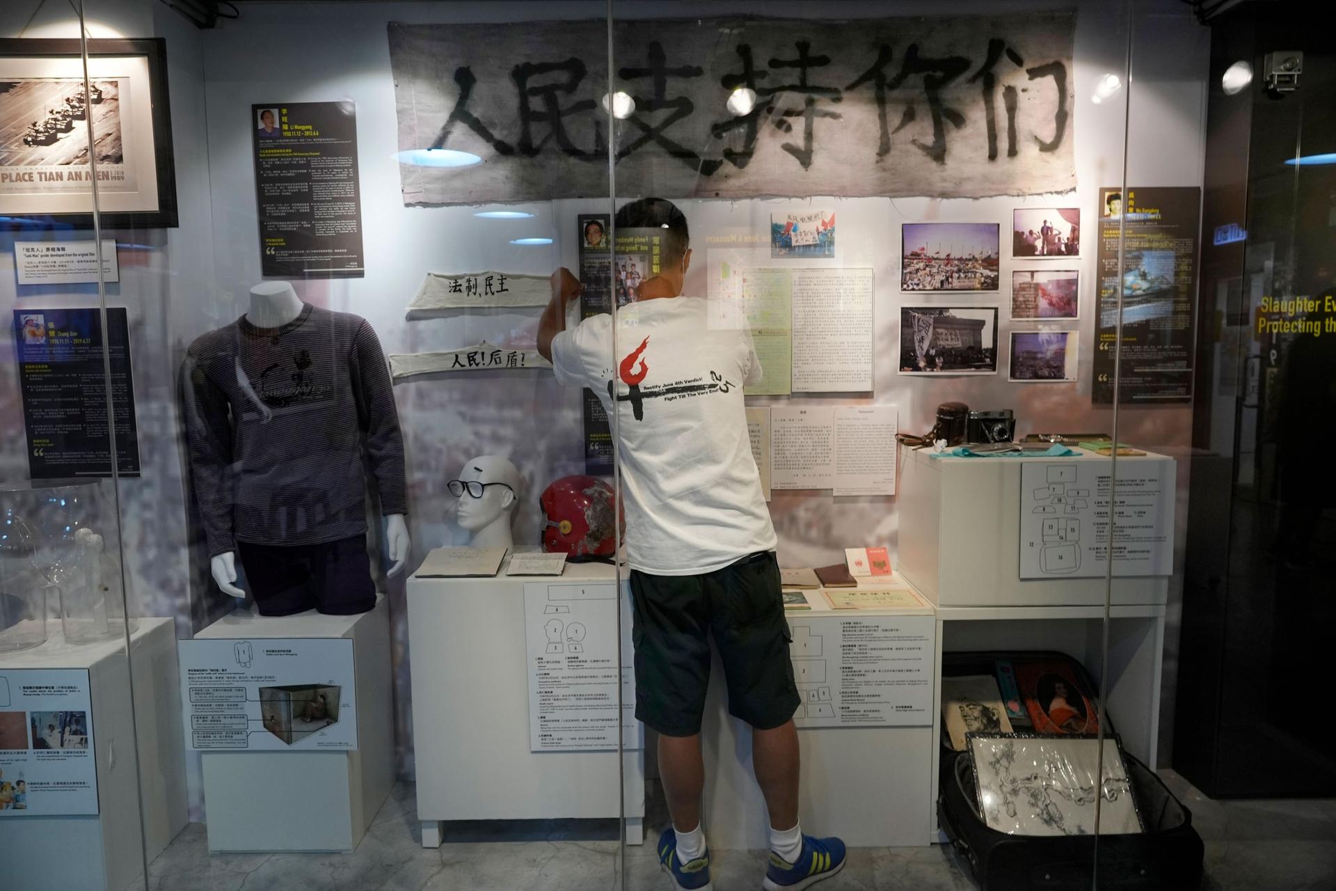 A man is shown wearing shorts and a t-shirt and standing in front of Tiananmen Square artifacts both hanging on the wall and on a mannequin. 
