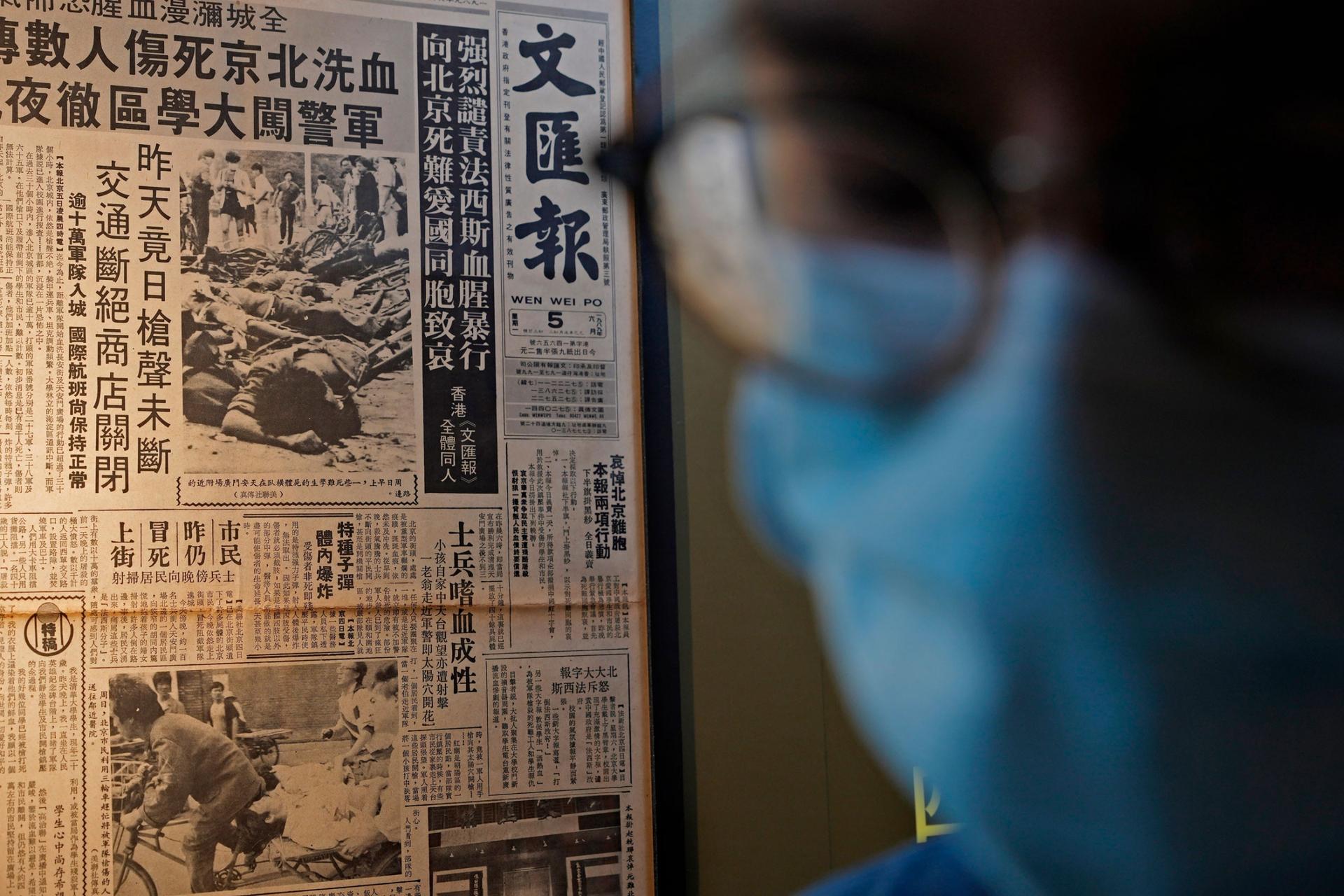 A newspaper clipping is shown in the background with a person in the near ground wearing a facemask and glasses is shown in blurred focus.