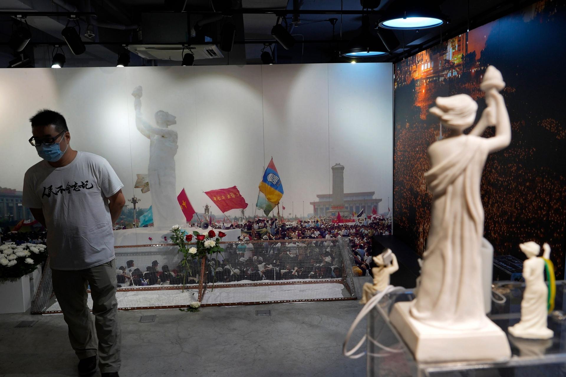 A man is shown wearing a white t-shirt, face mask and glasses while standing in front of a large wall-sized photograph depicting the Goddess of Democracy statue.