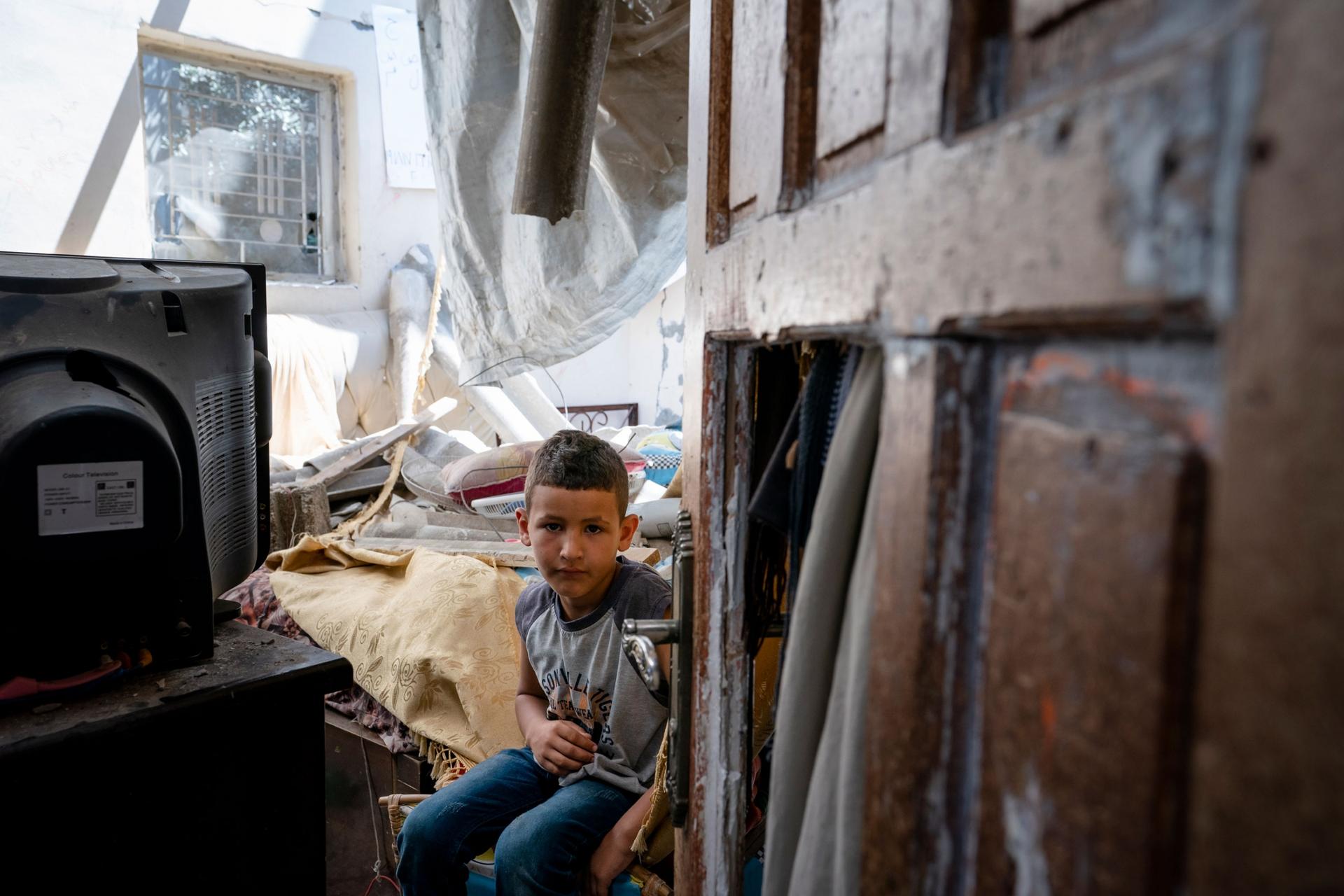 A young boy is shown seated next to a television and a damaged door in the near ground.