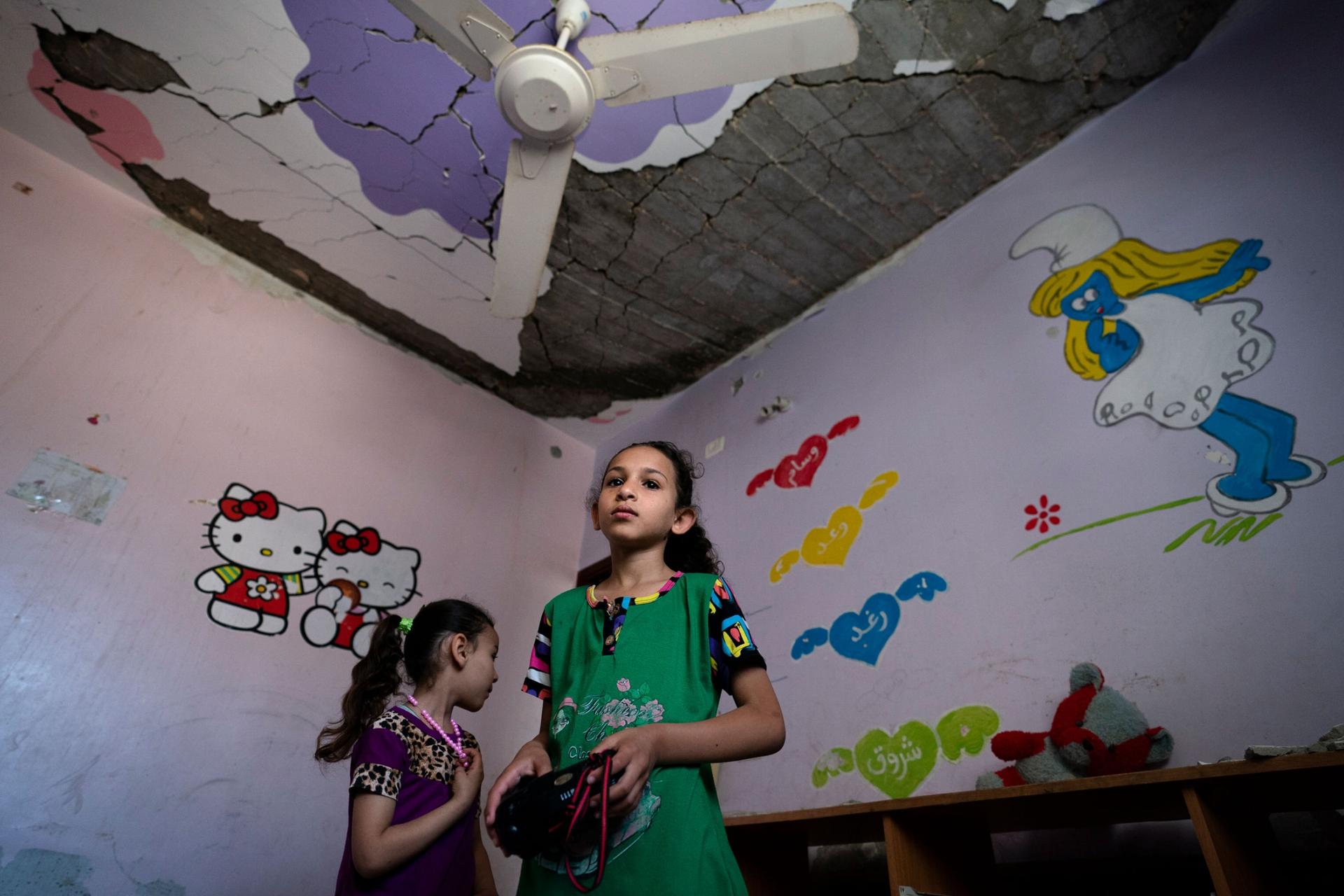 Two young girls are shown in a room with a smurf cartoon on the wall and a damaged ceiling.
