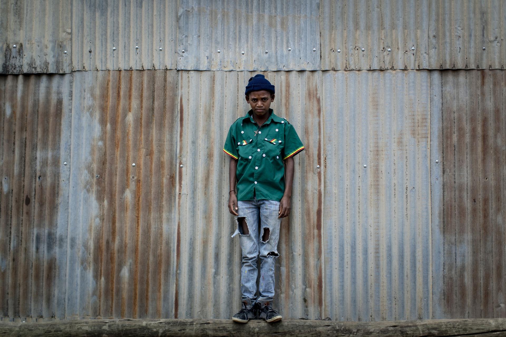 boy stands against corrugated steel wearing a green cultural shirt with buttons and jeans. 