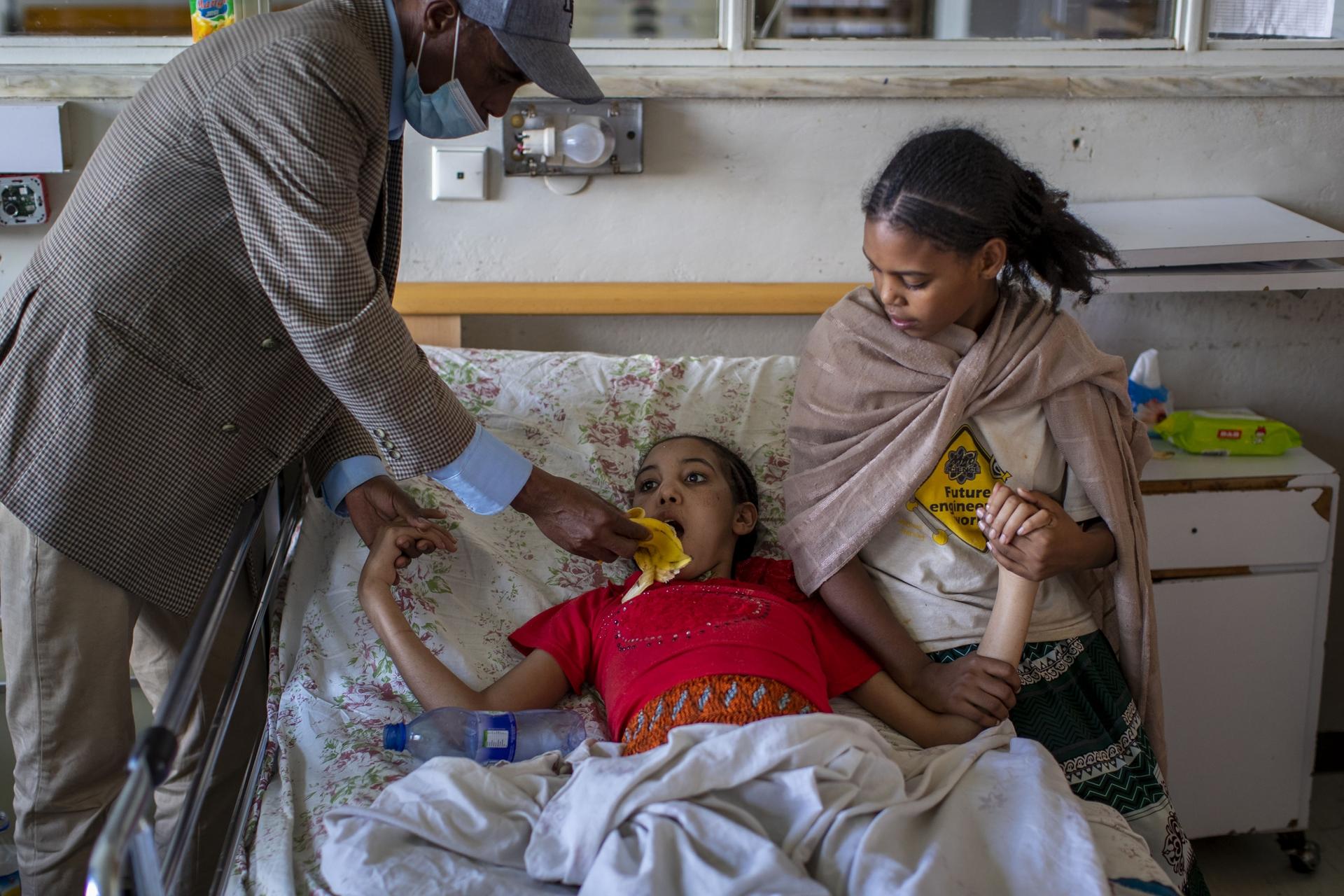 A girl in red rests on bed with her father and sister helping her. 
