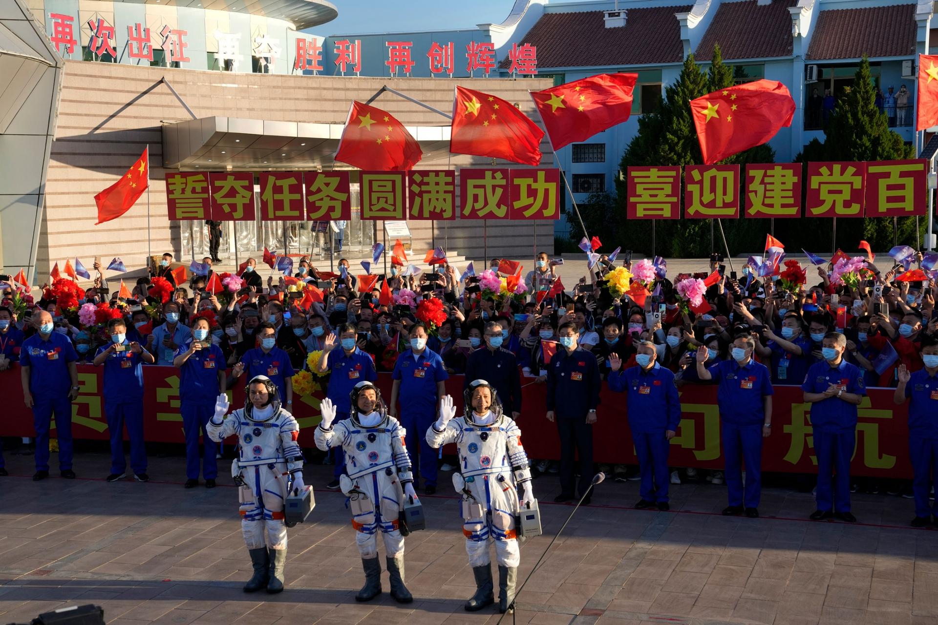 Three Chinese astronauts are shown waving with a large crowd of people waving the Chinese flag behind them.