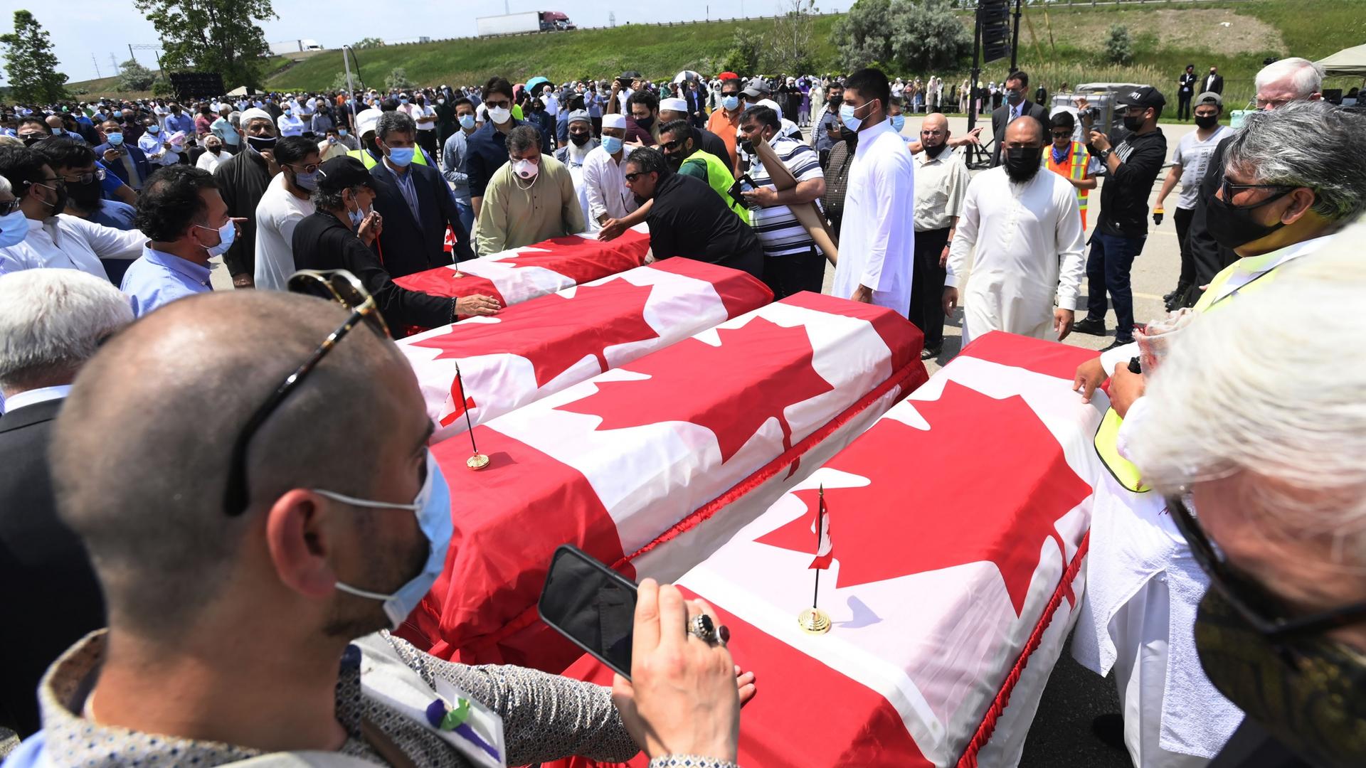 Four caskets are covered with the Canadian flag of red and white as mourners look on. 