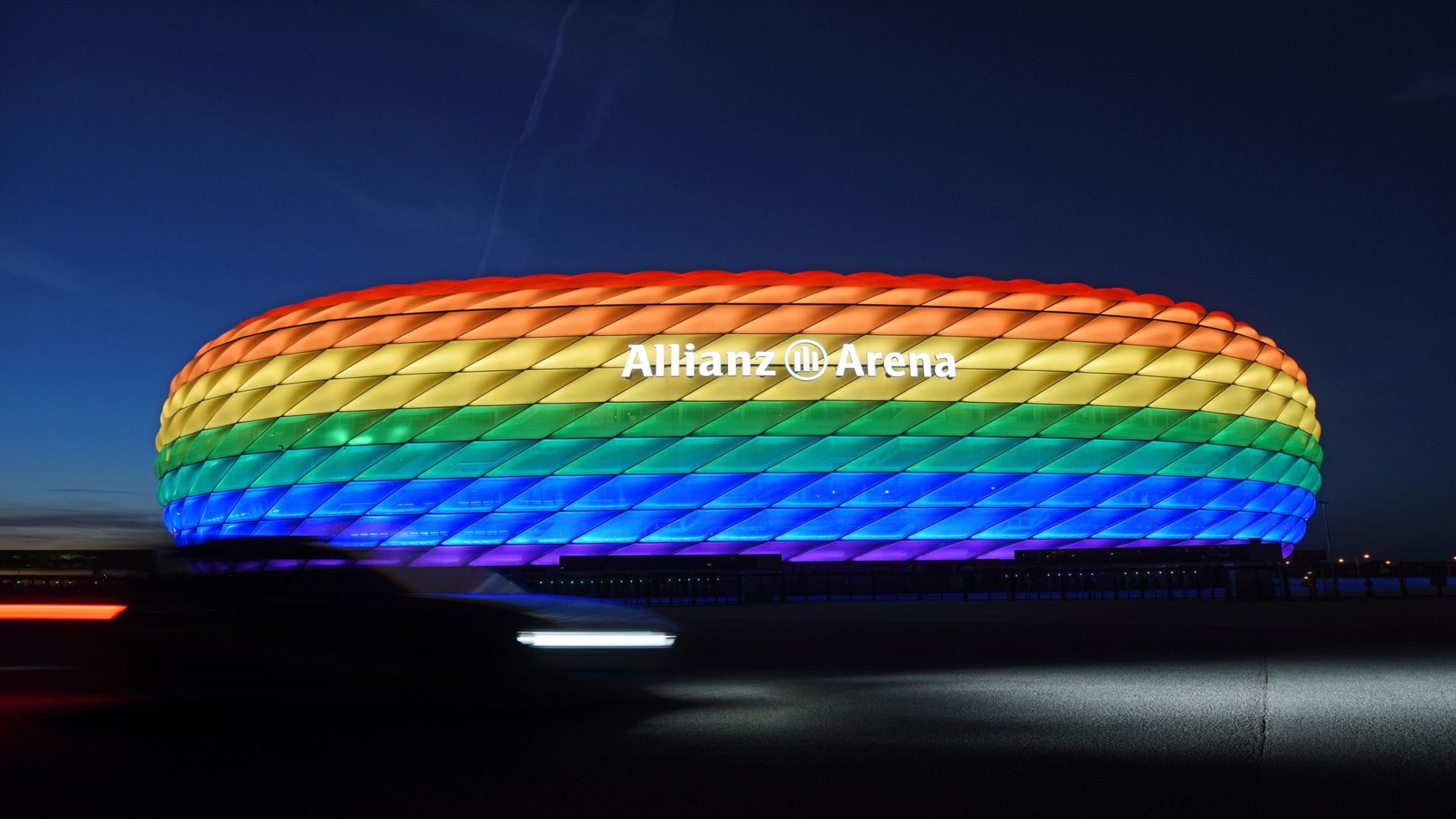 The Allianz Arean in Munich is shown illuminated in the colors of the rainbow with the night sky in the background.