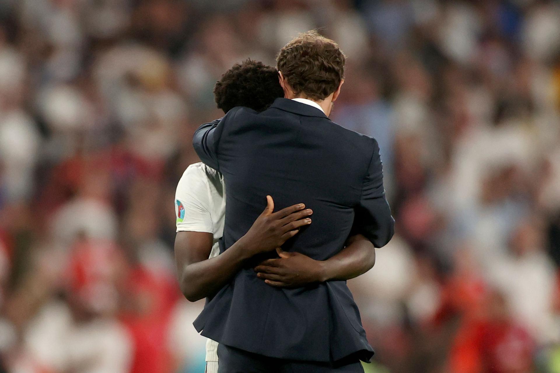 England's manager Gareth Southgate is shown hugging Bukayo Saka on the soccer pitch with fans shown in blurred focus in the distance.
