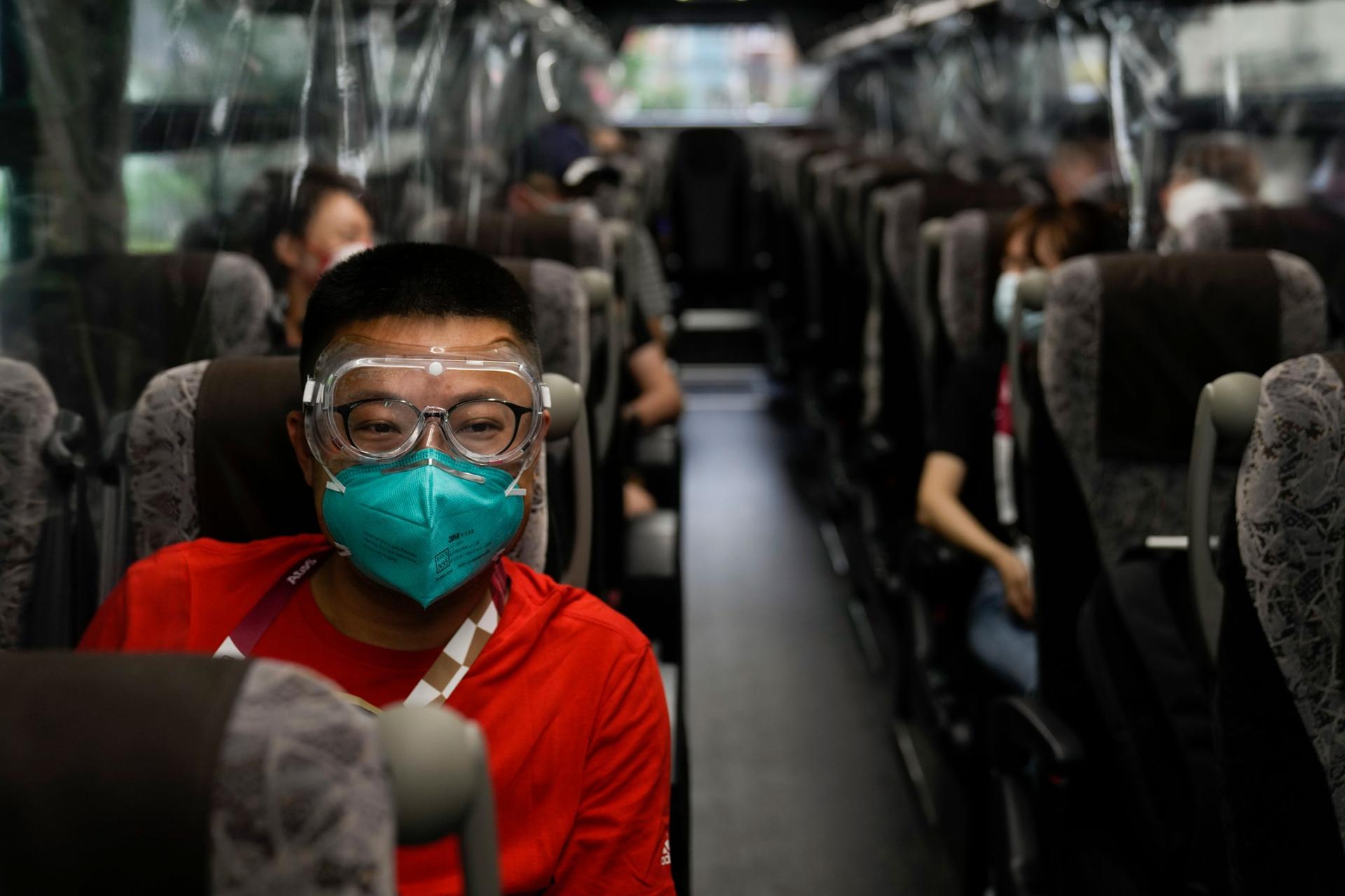A man is shown wearing a red shirt, face mask and clear goggles while riding a bus.