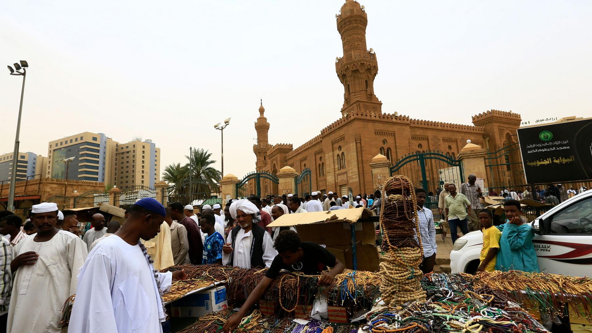 A large crowd od people are shown outside of the tan-colored Grand Mosque with a man selling beaded jewelry in the nearground.