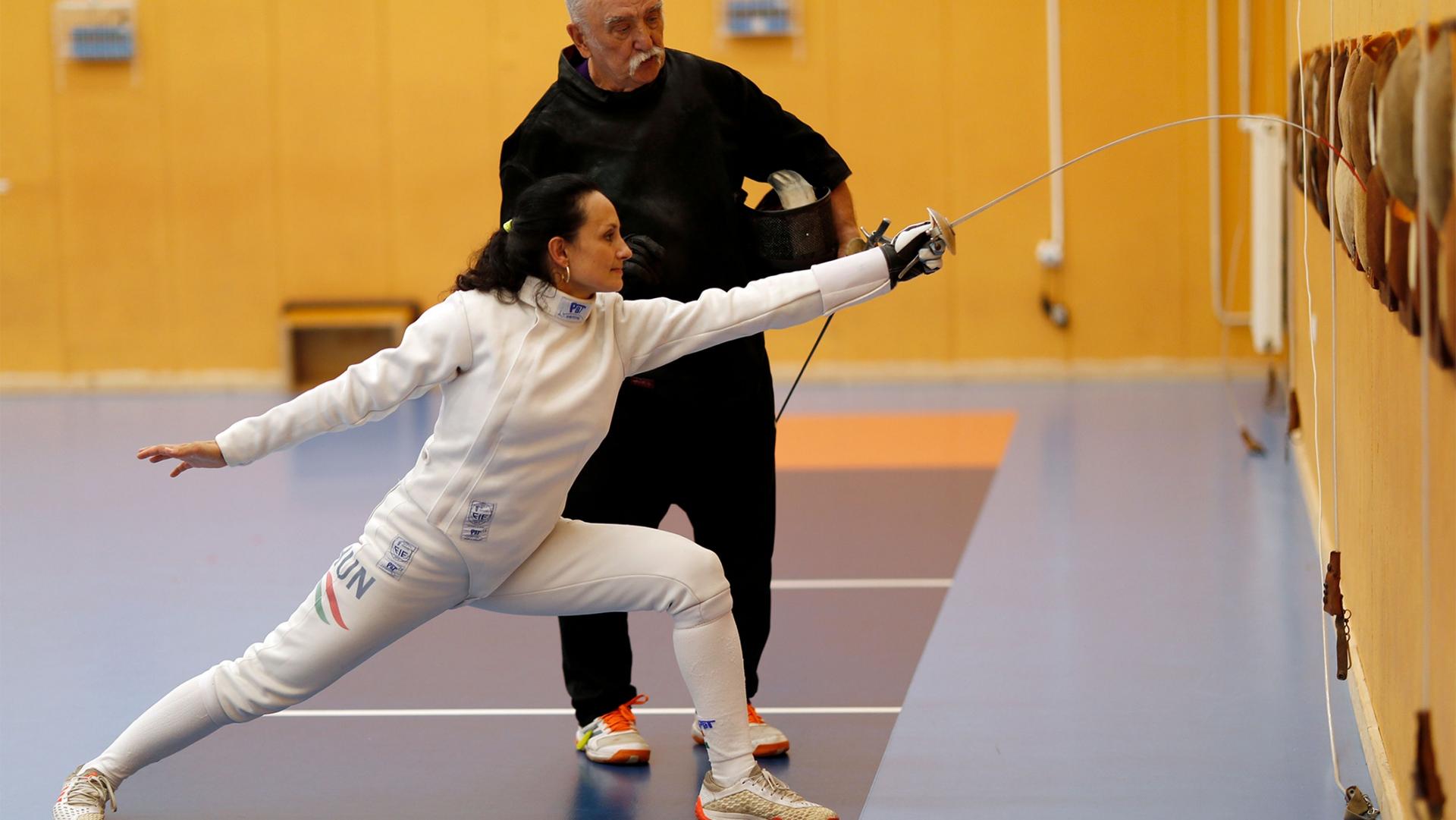 Woman in white fencing gear trains with her coach standing behind her wearing black