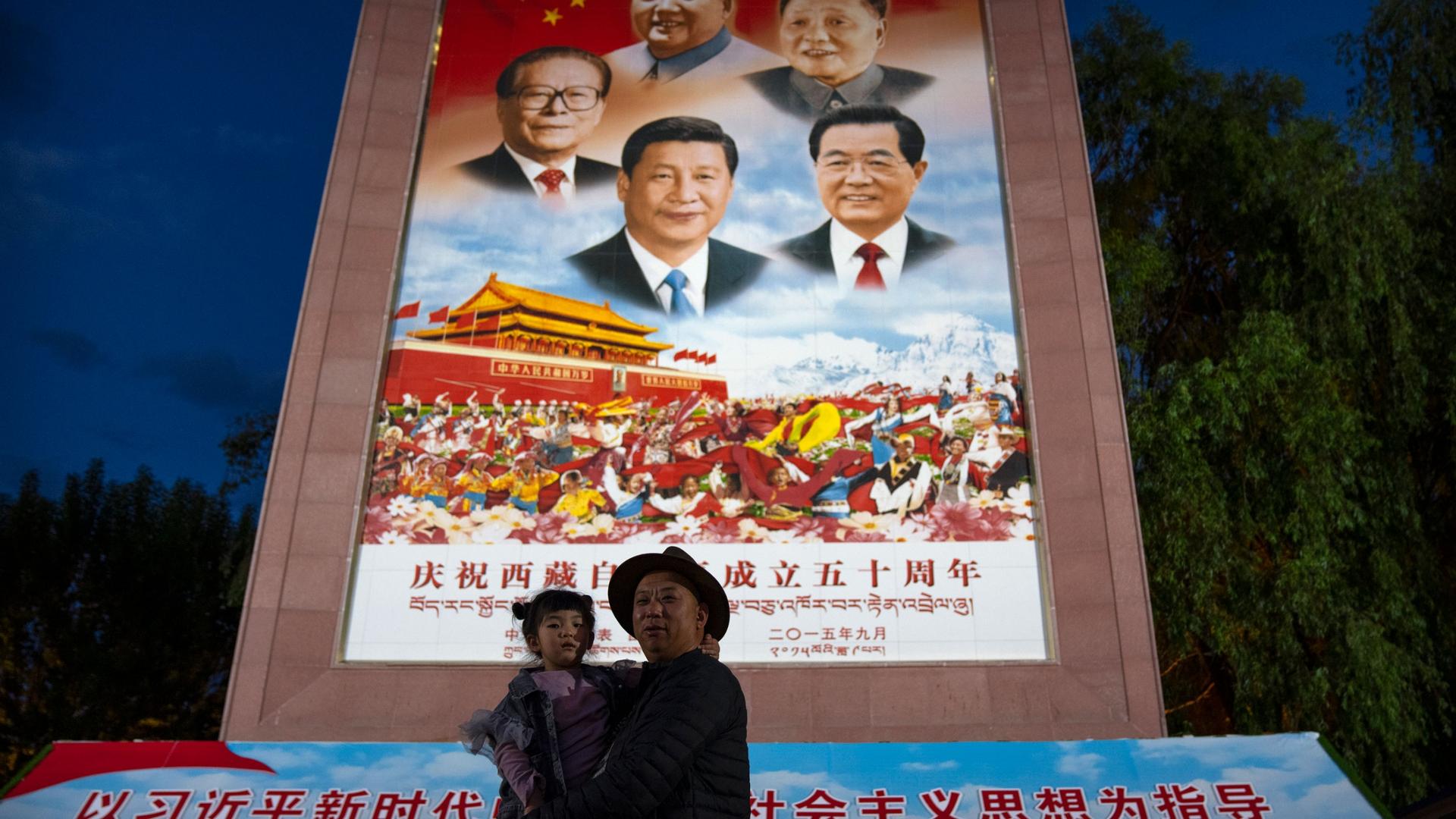 A man holds a girl as they pose for a photo in front of a large mural depicting Chinese President Xi Jinping, bottom center, and other Chinese leaders at a public square at the base of the Potala Palace in Lhasa in western China's Tibet Autonomous Region 