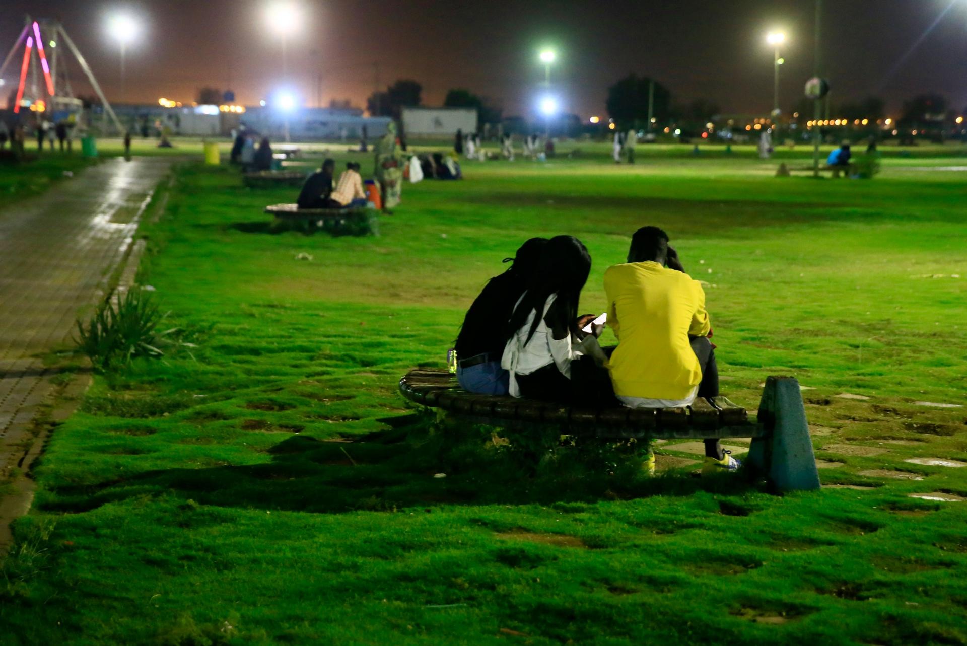 Three people are shown with one person wearing a yellow shirt while sitting on a curved metal bench amid a large grassy area.